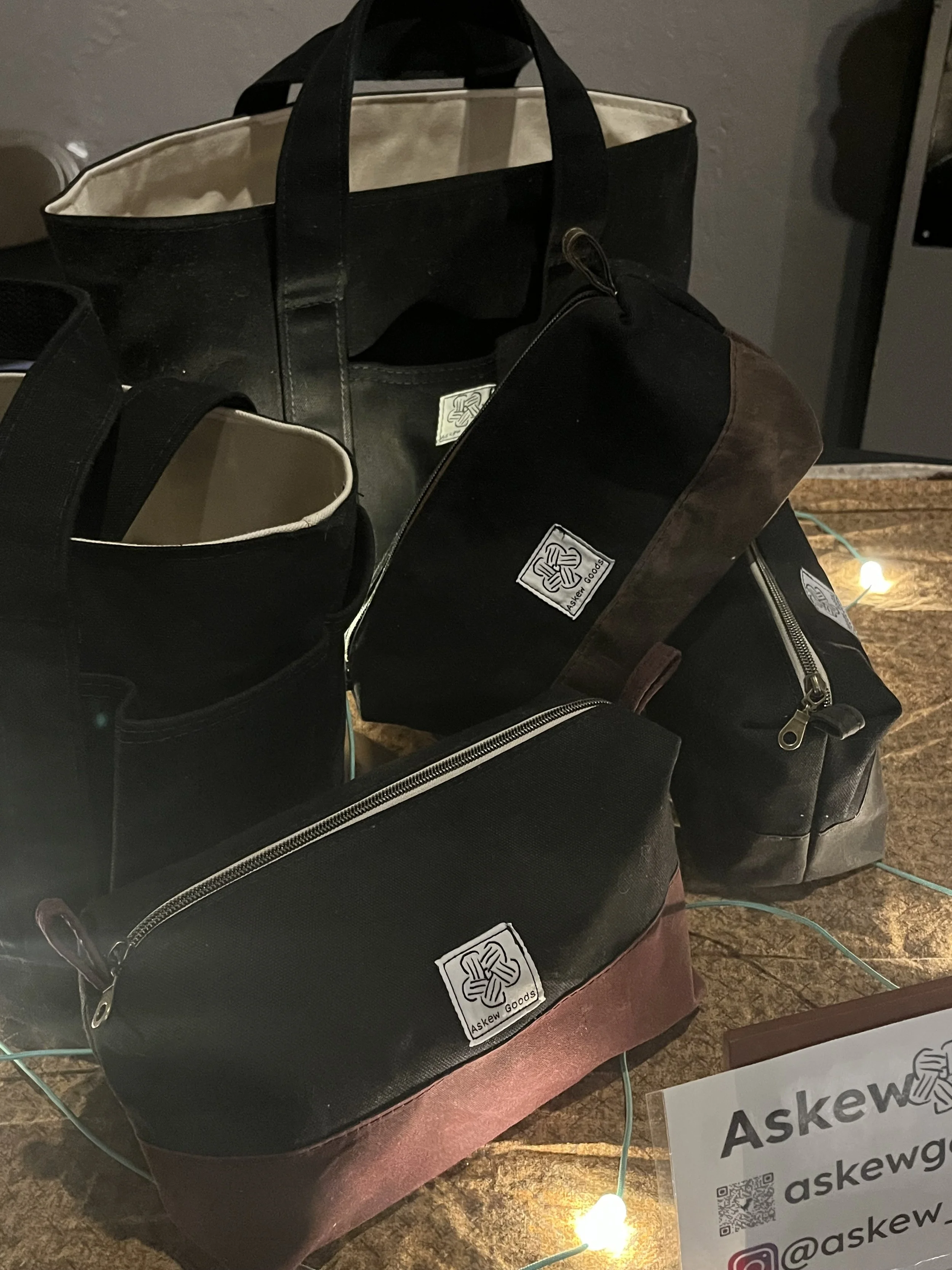 Black and brown bags on a wooden surface with a string of small lights and a sign in front with the text 'Askew' and social media handles.