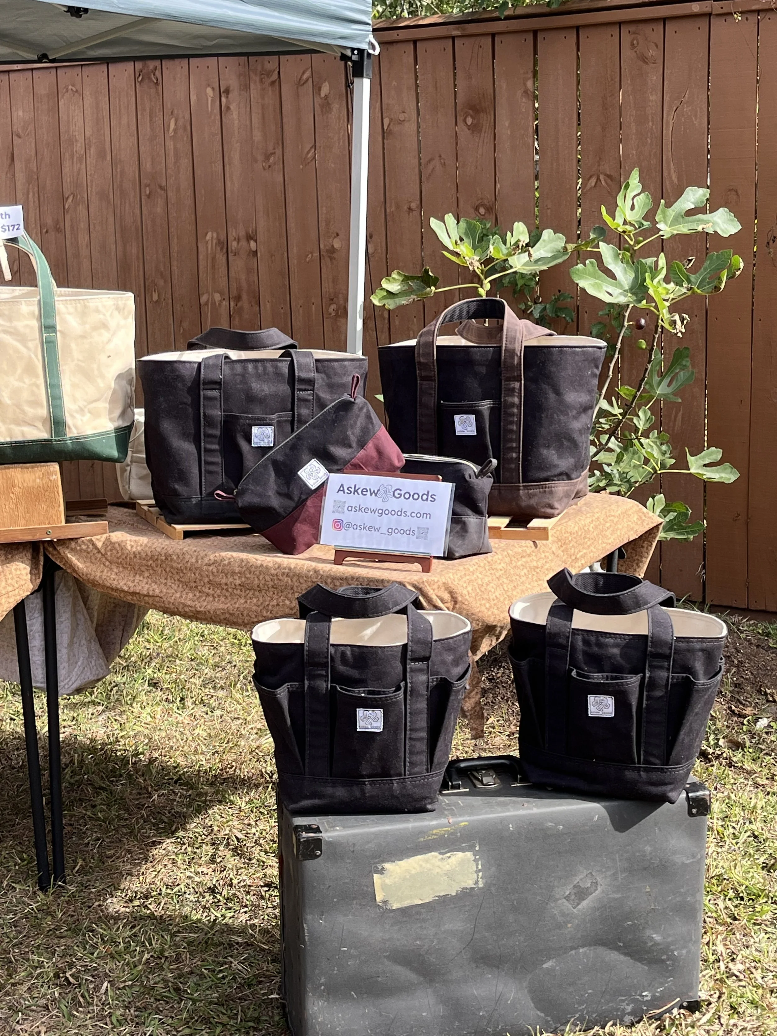 Display of various black and beige bags and containers at an outdoor market stall, with a wooden fence and green plants in the background.
