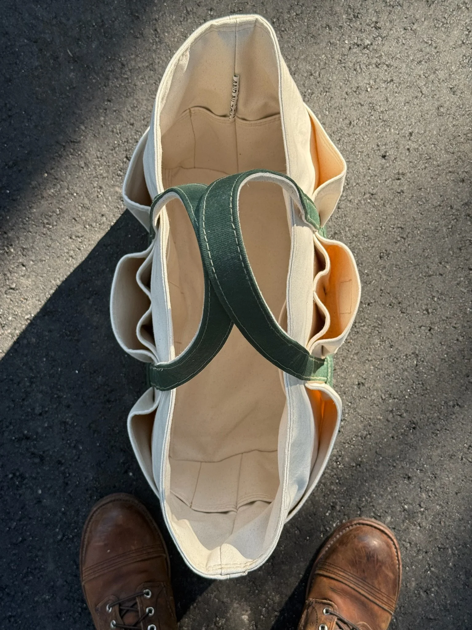 A top-down view of an empty canvas tote bag with green handles on a dark asphalt surface, with a person's brown leather boots visible at the bottom of the frame.