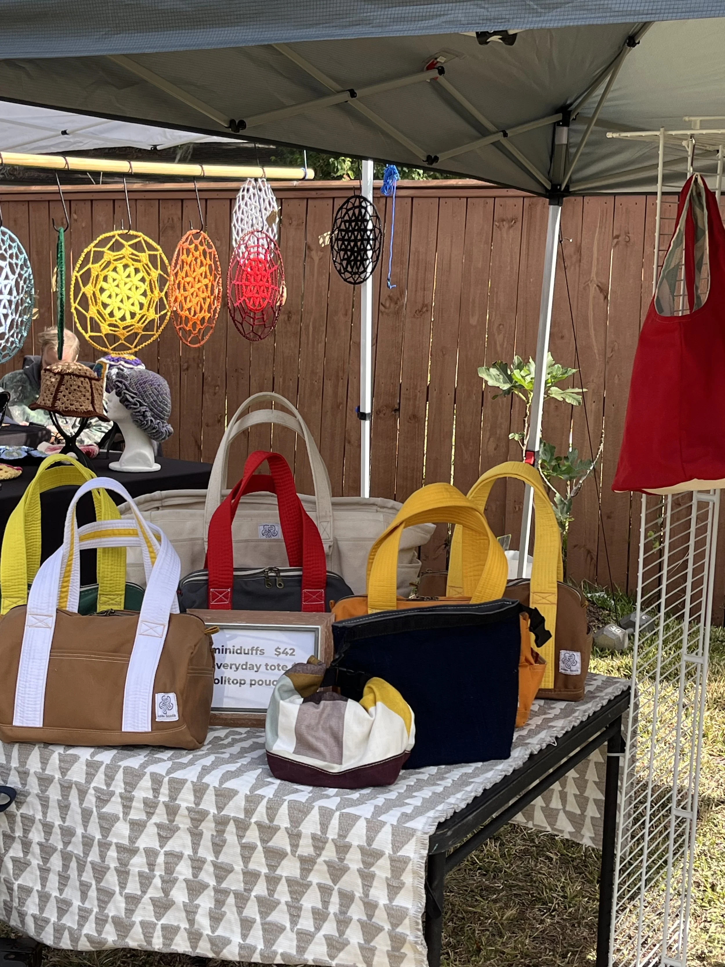 A market stall selling colorful crochet bags and hats, with a variety of bags in brown, yellow, white, red, and striped patterns arranged on a table. Decorative crochet ornaments hang above, and a wooden fence and greenery are in the background.