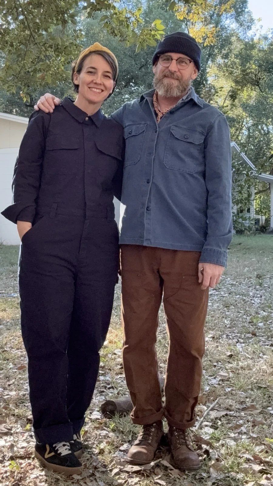 A smiling woman and a man with a beard and glasses standing outdoors, arm around each other, in a backyard with trees and a house in the background.
