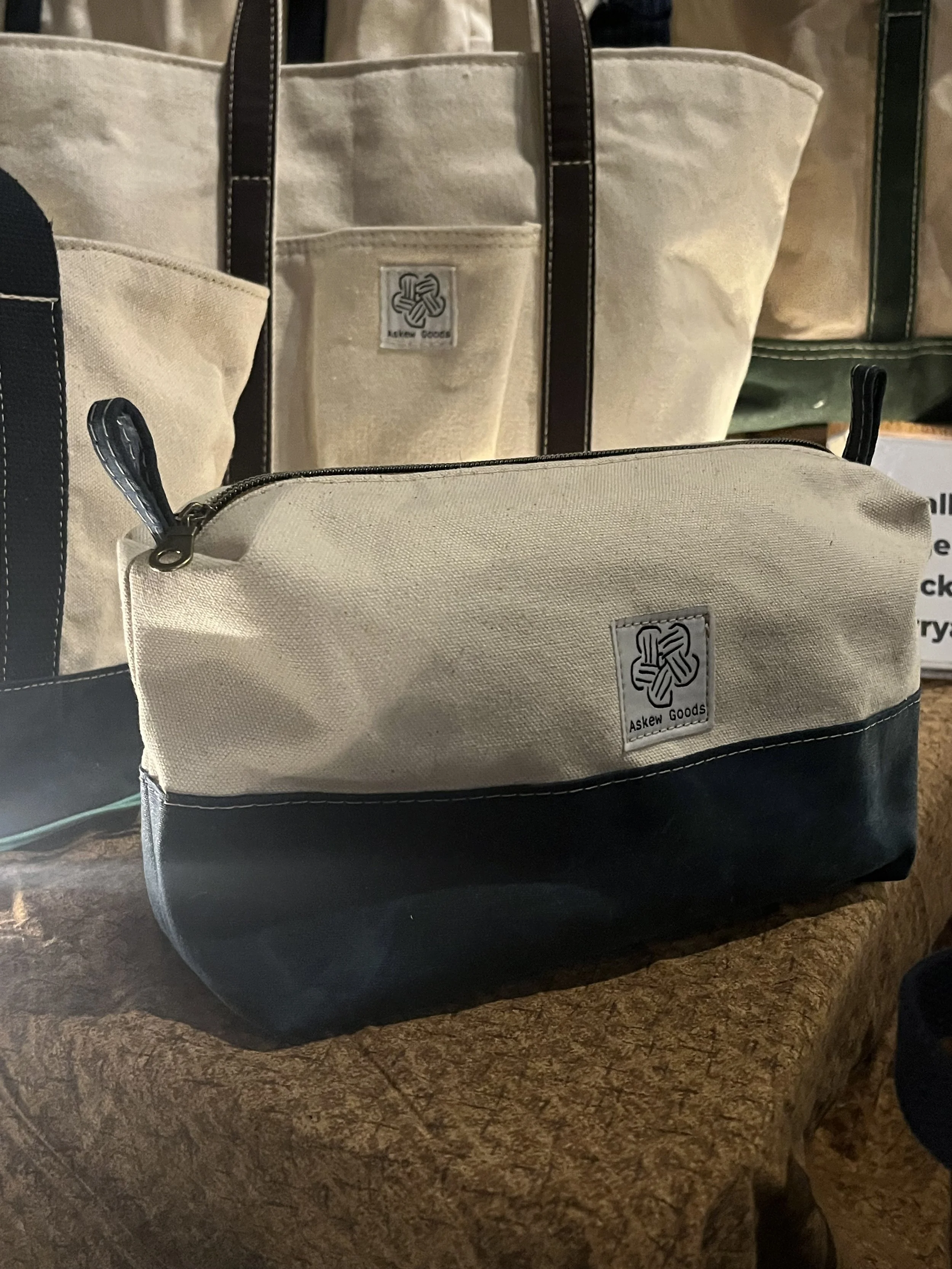 Close-up of a beige and black layered tote bag with a zipper compartment, placed on a textured brown surface. The bag has a logo patch that reads 'Askew Goods' with a knot symbol.