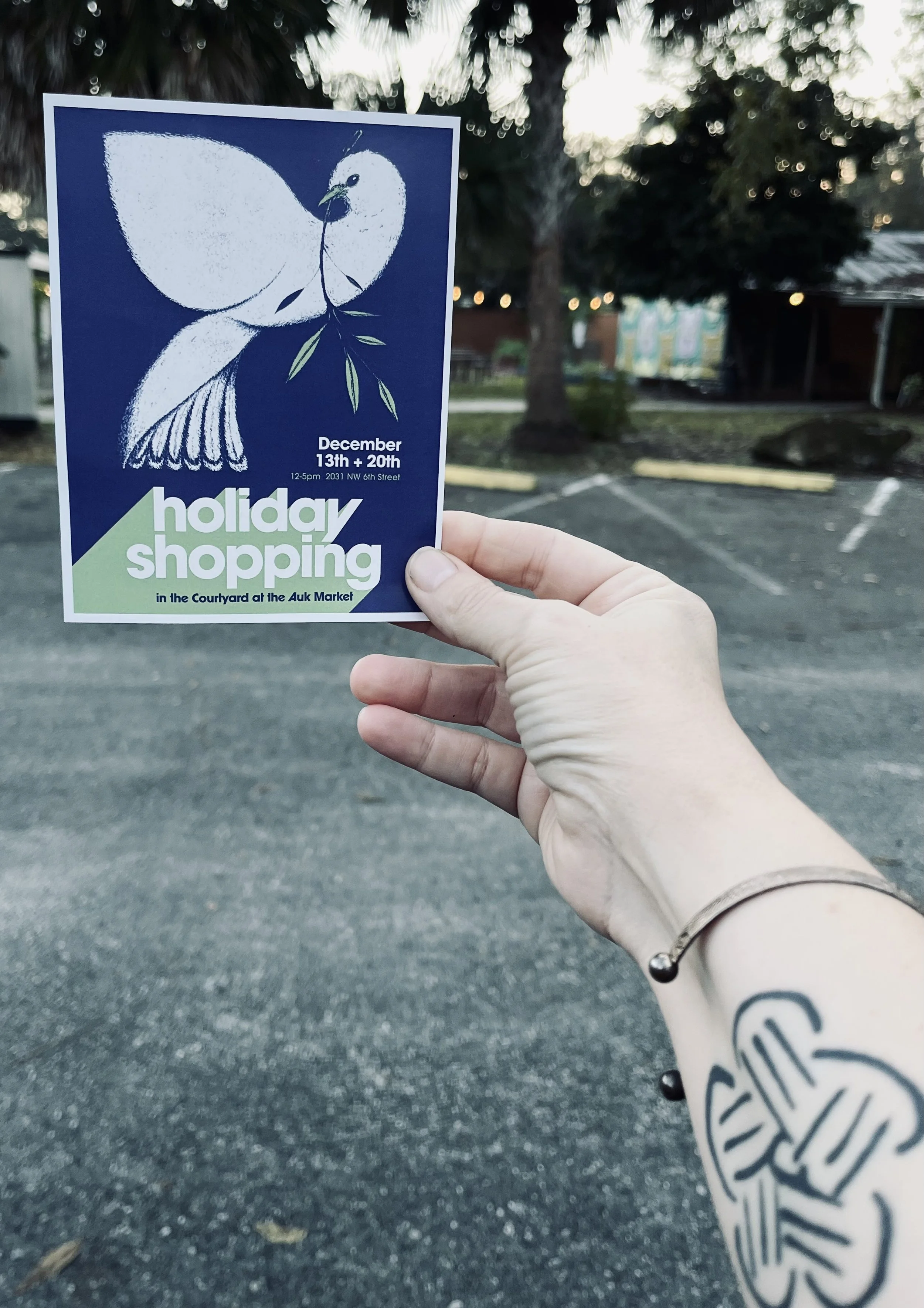 A person holding a flyer that advertises a holiday shopping event at the Auk Market, featuring a white dove illustration on a blue background, with details about the event date and time, in an outdoor parking lot.