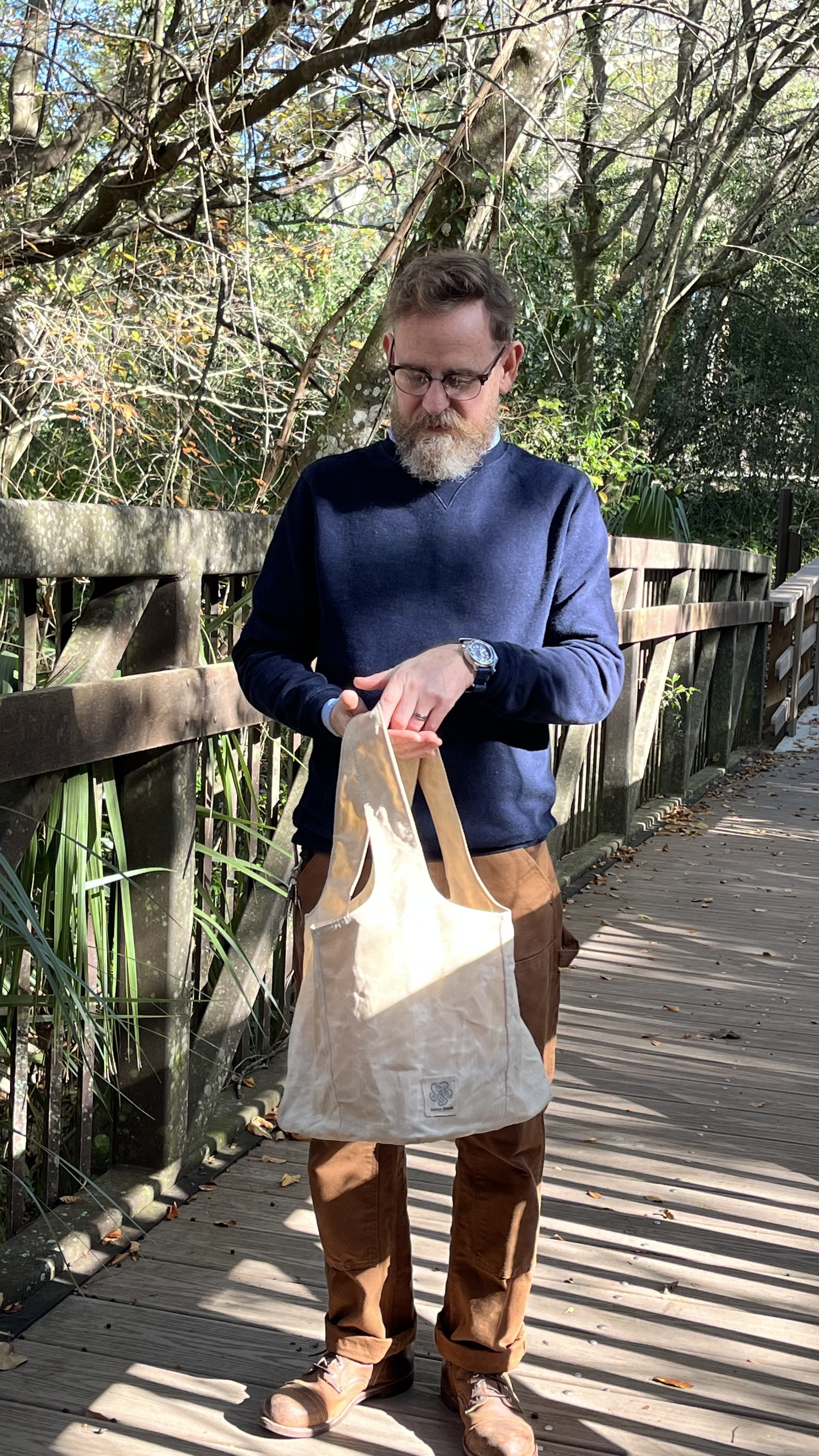 A man with glasses and a beard, wearing a navy sweater and brown pants, is standing on a wooden walkway surrounded by trees and foliage. He is holding a large beige tote bag.