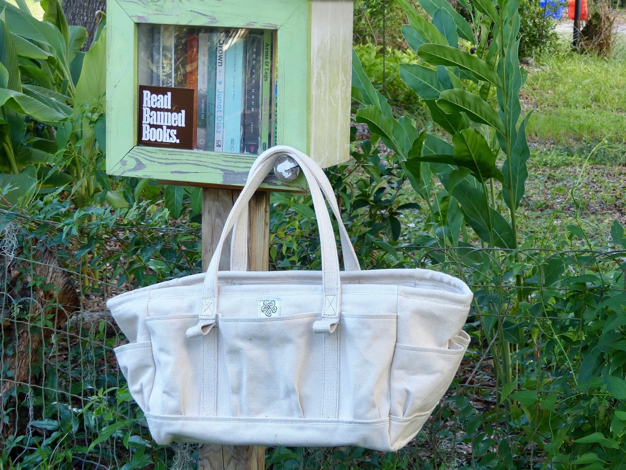 A wooden mailbox with a glass window holding books, mounted on a wooden post, surrounded by green foliage and a white tote bag hanging from the post.