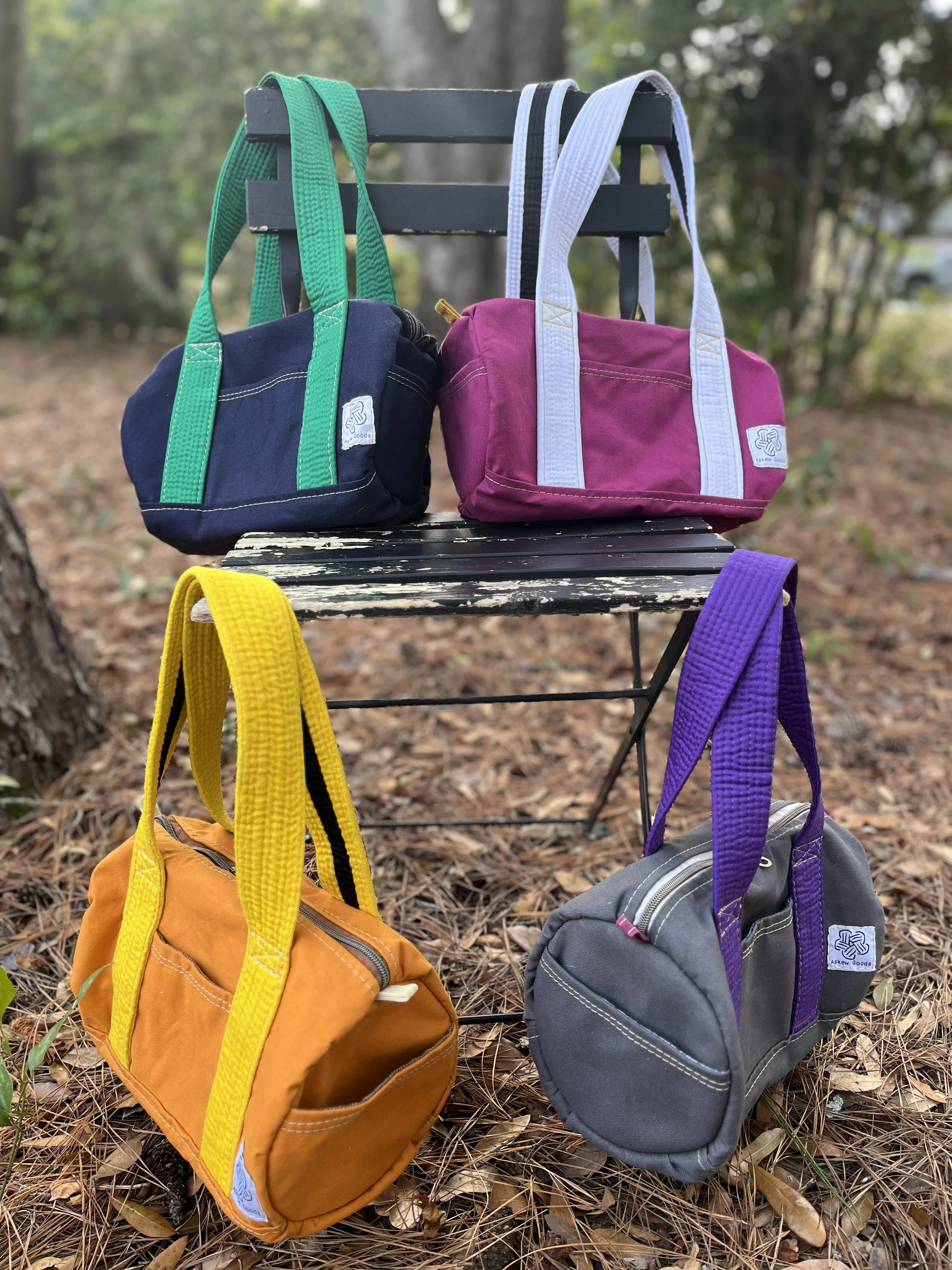 Four colorful backpacks hanging from a black metal outdoor chair on a ground covered with pine needles, with a wooded background.