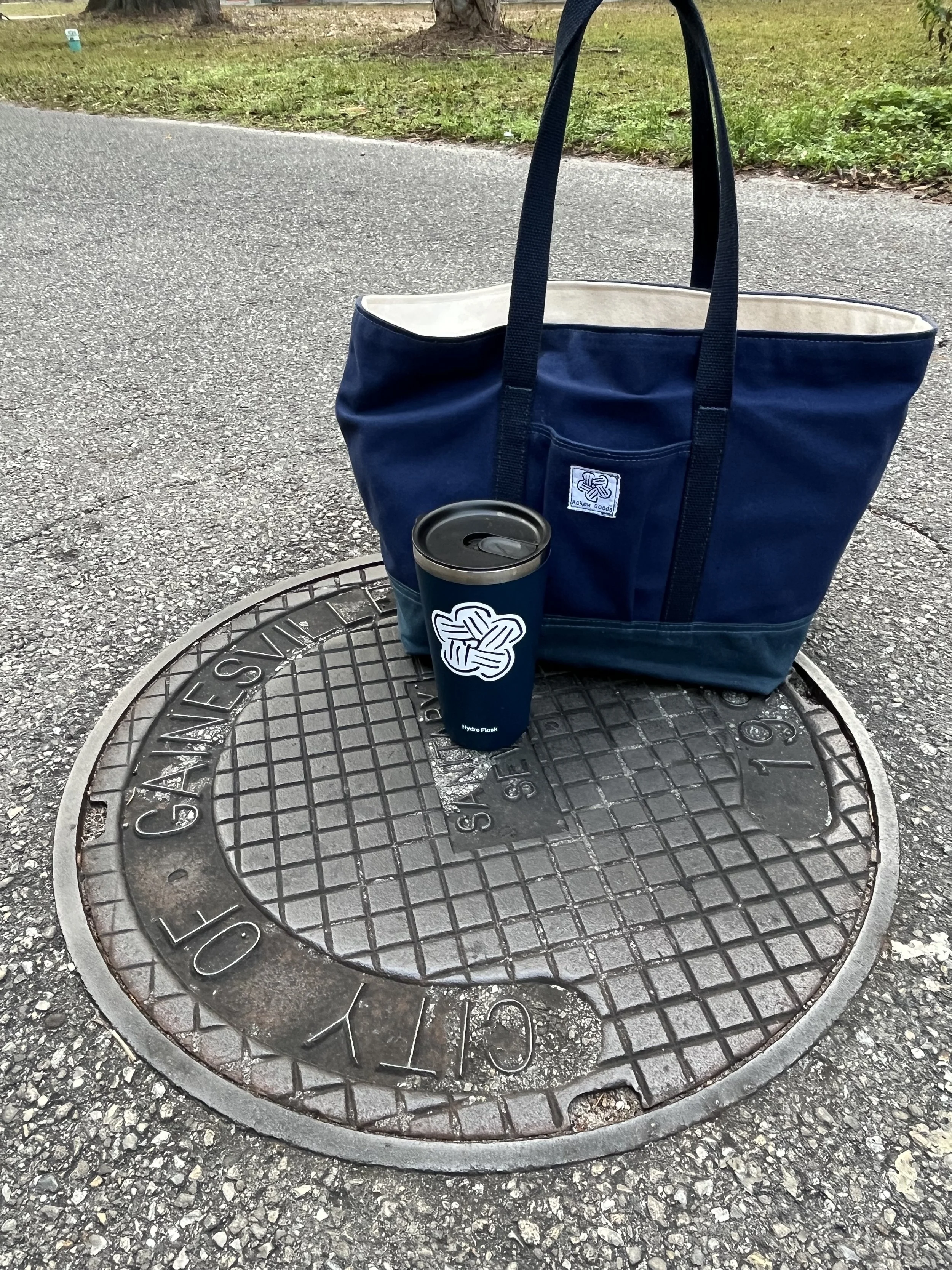 A blue tote bag and a reusable coffee cup placed on a manhole cover on a paved surface, with grass and some trees in the background.