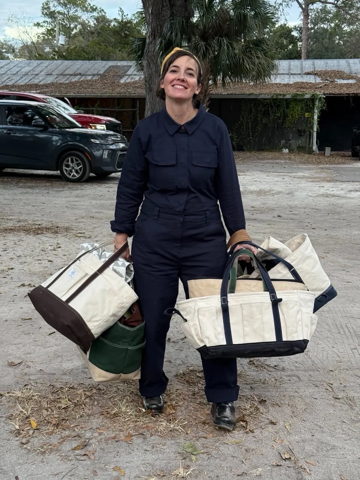 Woman wearing a navy jumpsuit and yellow headband smiling outdoors, carrying multiple large bags filled with various items, standing on dirt ground with trees, parked cars, and a weathered building in the background.