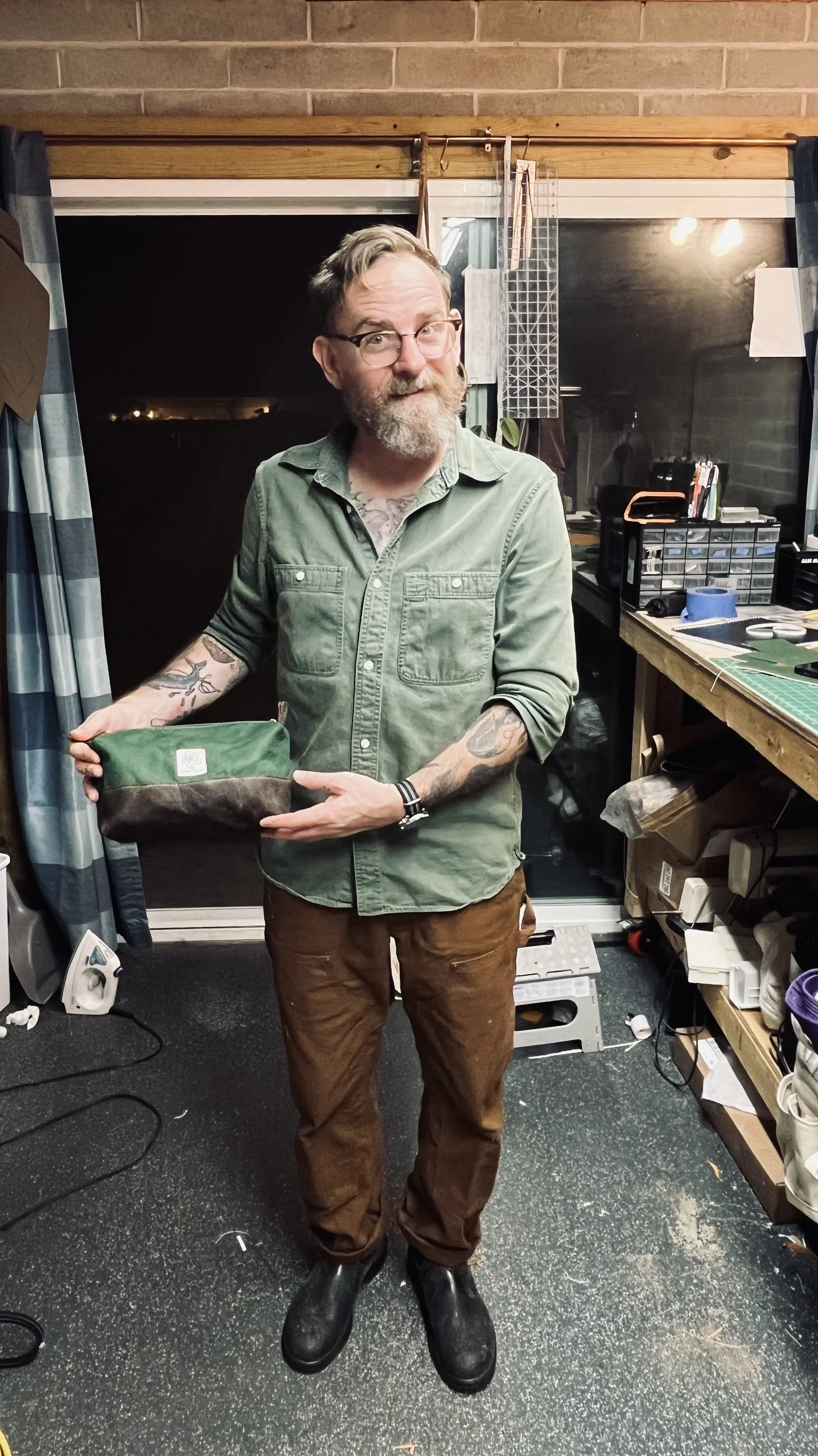A man with glasses and a beard, wearing a green button-up shirt and brown pants, is standing in a cluttered room at night, holding a small fabric item. The room has shelves, boxes, and various objects, with a window behind him showing darkness.