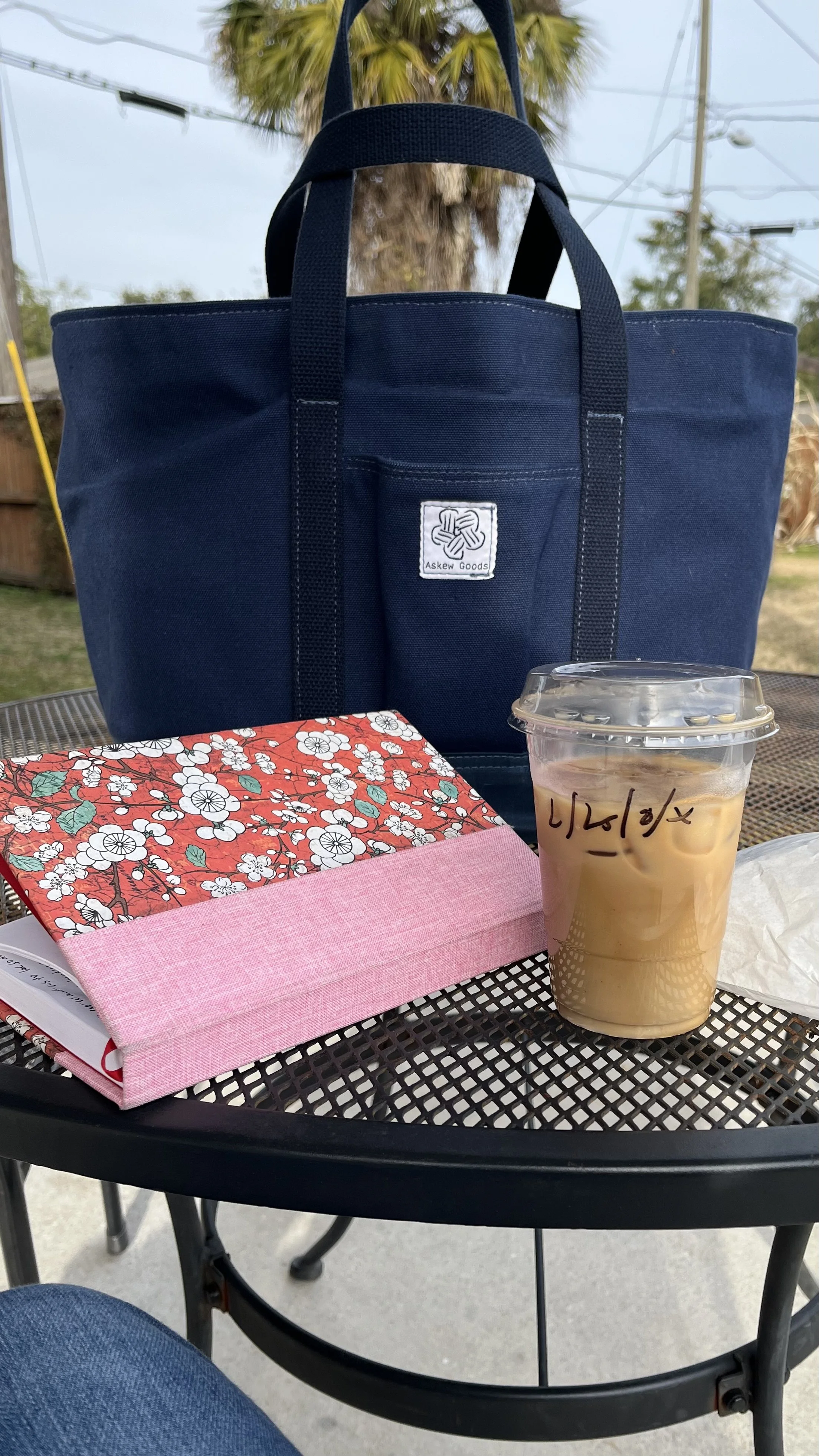 A black metal outdoor table with a pink box, a floral-patterned fabric-covered book, and a plastic cup of iced coffee with writing on it. In the background, there is a navy blue tote bag with a logo, a palm tree, and power lines.