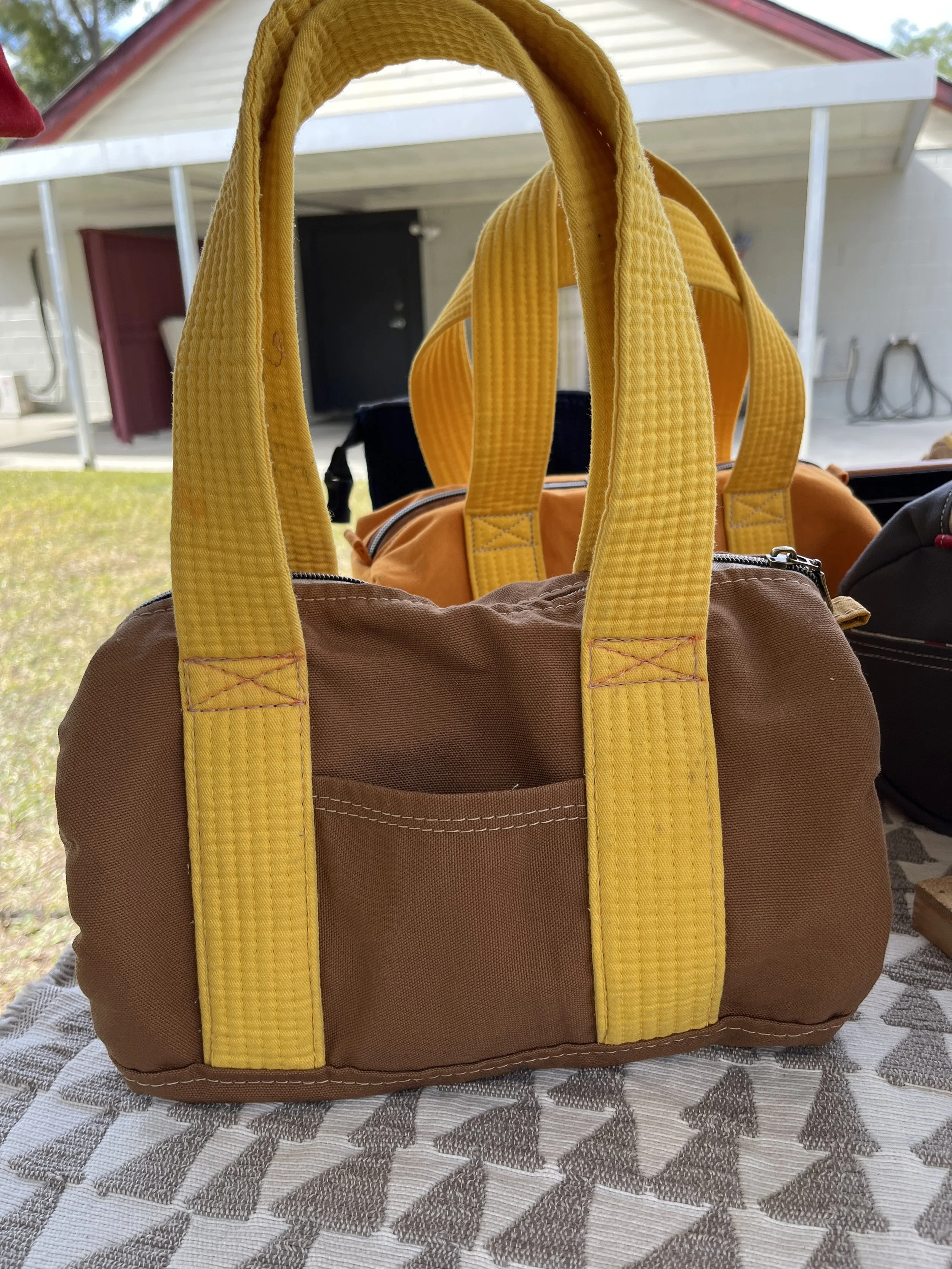 Brown and yellow bag with yellow straps placed on a patterned tablecloth, with a house and outdoor structure in the background.
