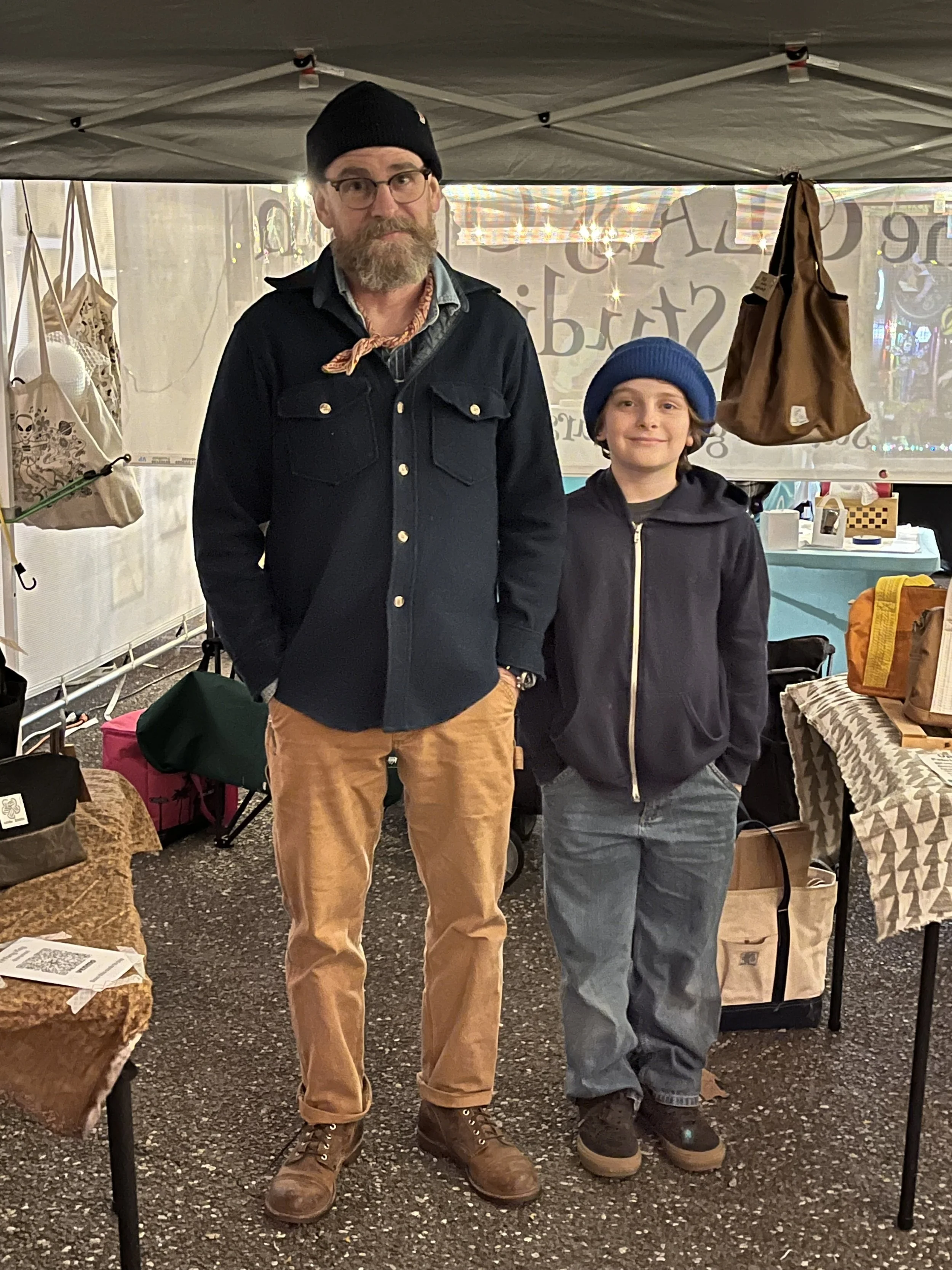 A man with glasses, a beard, and wearing a black beanie, denim jacket, and tan pants, standing next to a smiling boy with a blue beanie, gray hoodie, and gray pants, inside a tent at a market or fair with various bags and items on tables around them.