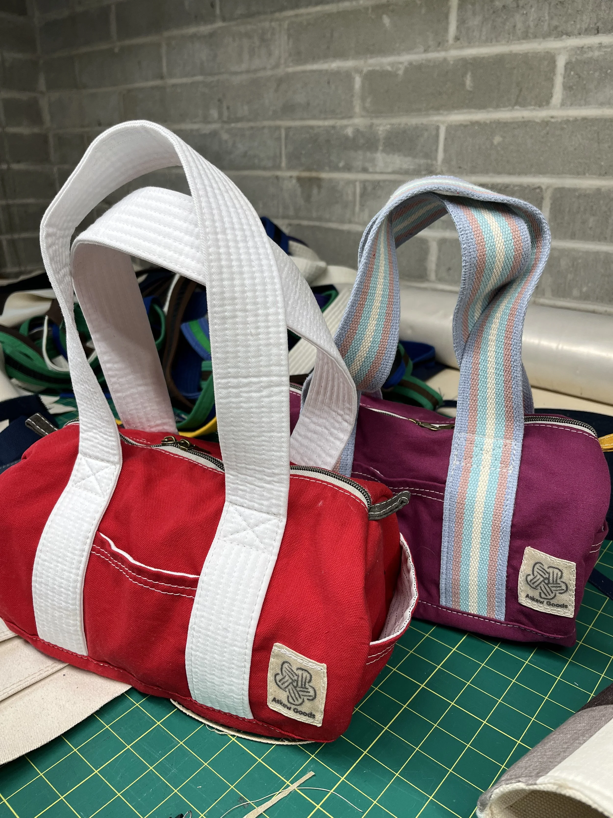 Colorful bags with striped handles, one red, and one purple, are on a green cutting mat with a grid pattern. A gray brick wall is in the background.