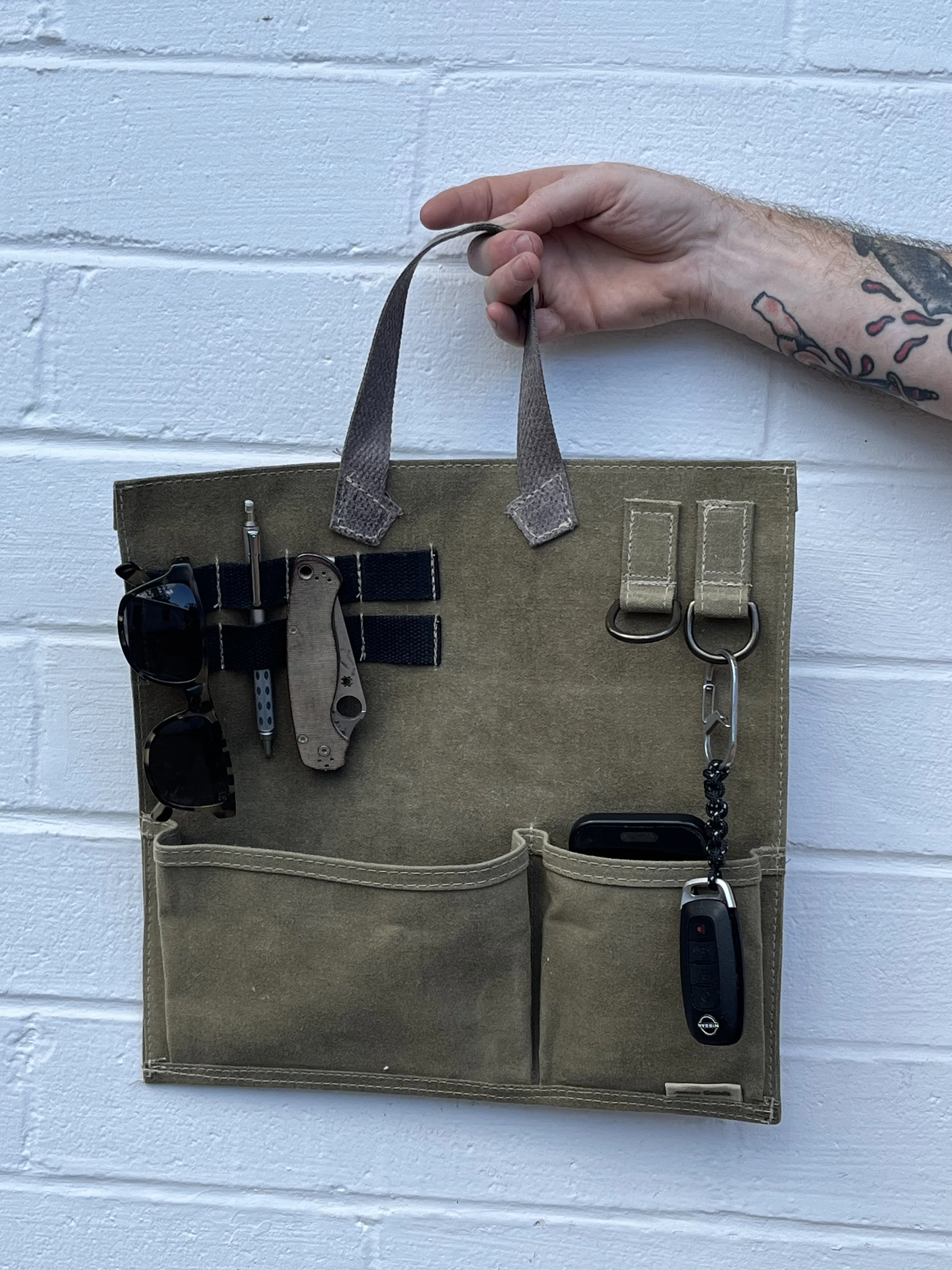 A person with a tattooed arm holding a beige organizer with various tools and accessories attached, including sunglasses, a keychain, and car keys, against a white brick wall.
