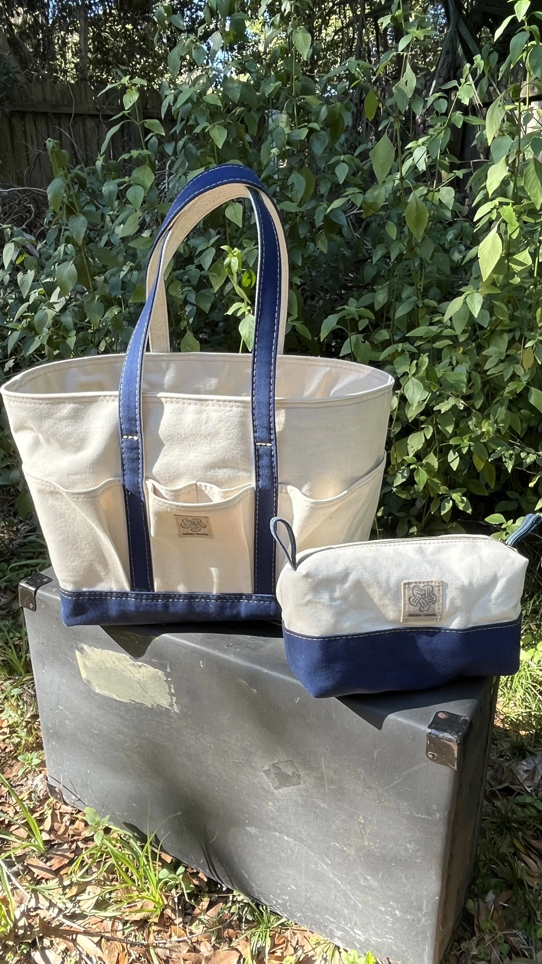 A collection of beige and navy tote bags and pouches with a logo, resting on a black surface outdoors with a background of green leafy bushes.