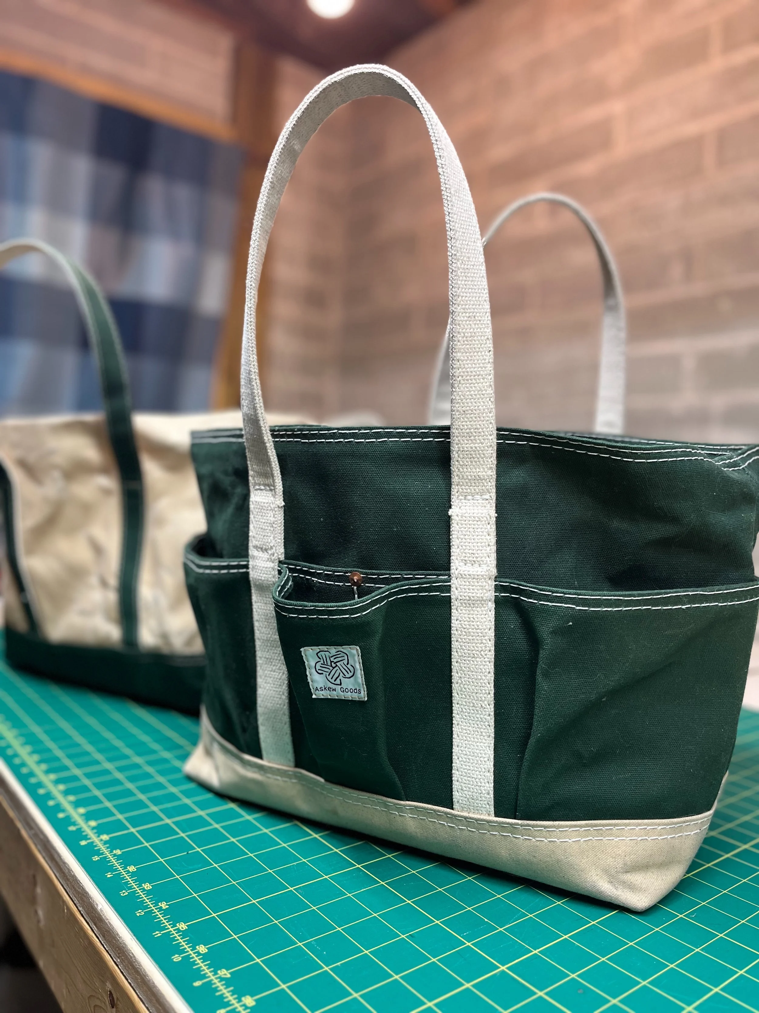 A black and gray tote bag with white straps and a small label that reads 'Askew Goods' sitting on a green cutting mat with yellow measurement lines in a workshop or craft space.