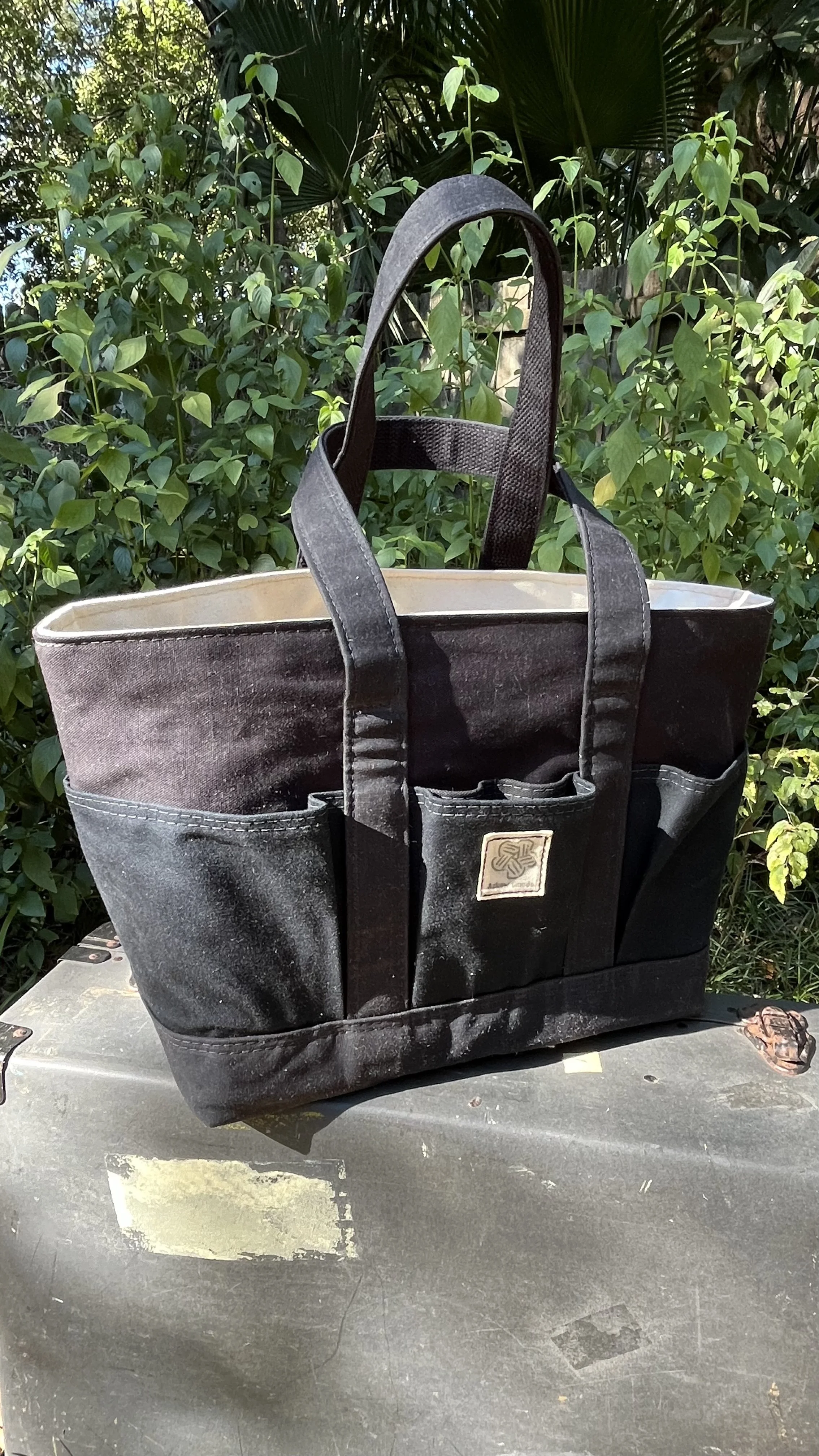 A black tote bag made of a durable, canvas-like material sitting outdoors on a gray surface with greenery in the background.