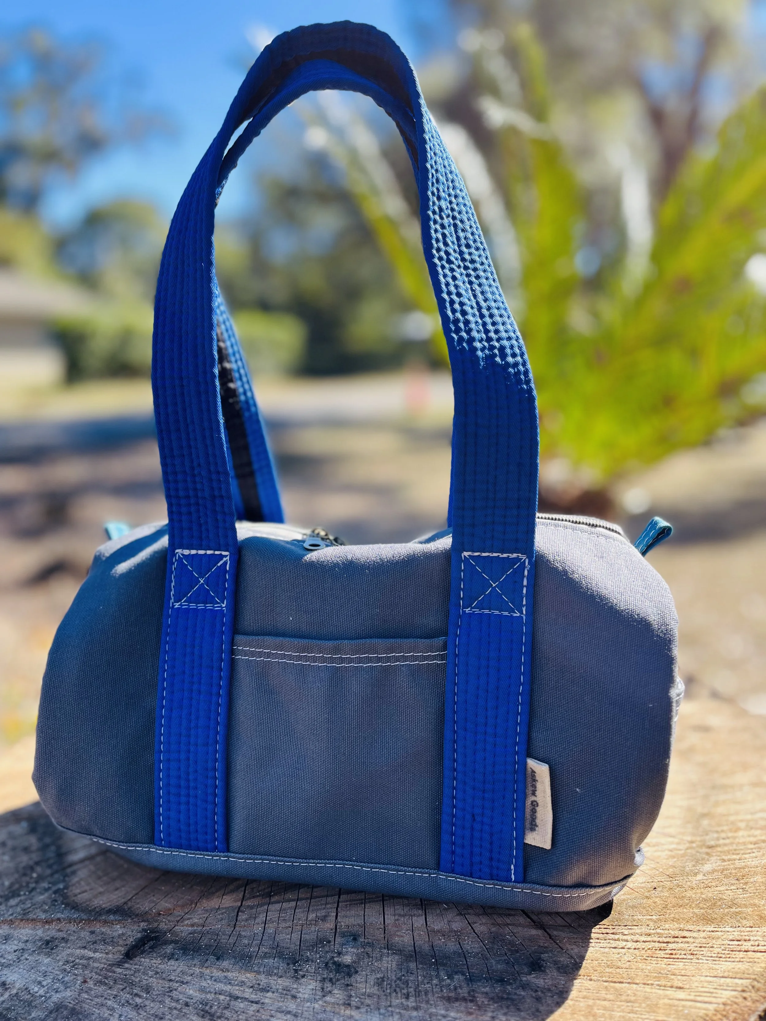 A gray backpack with blue straps sits on a wooden surface outdoors with trees and a blue sky in the background.