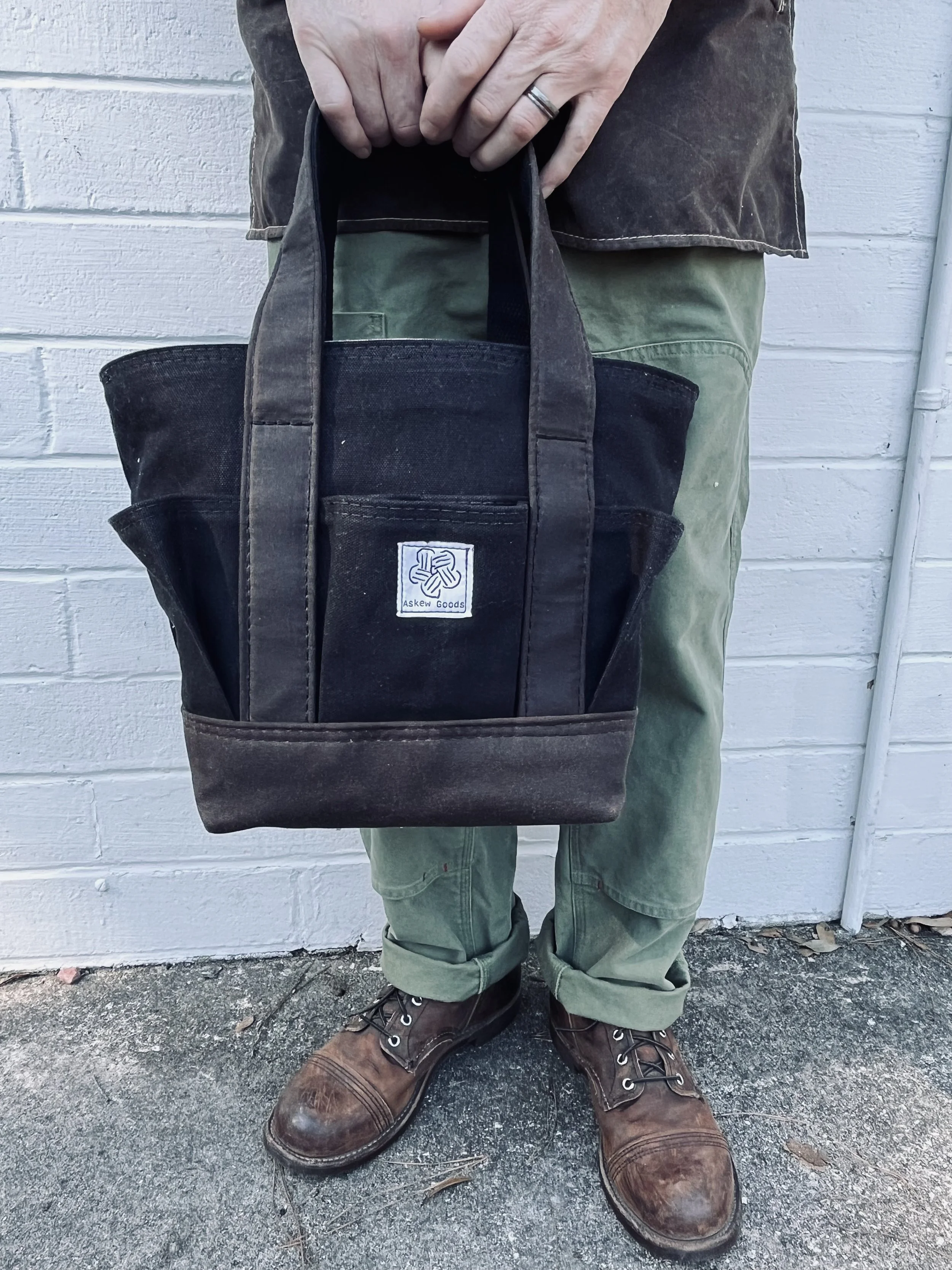 Person wearing brown leather boots, green pants, a brown shirt, and holding a black bag with a white logo, standing in front of a white wall.
