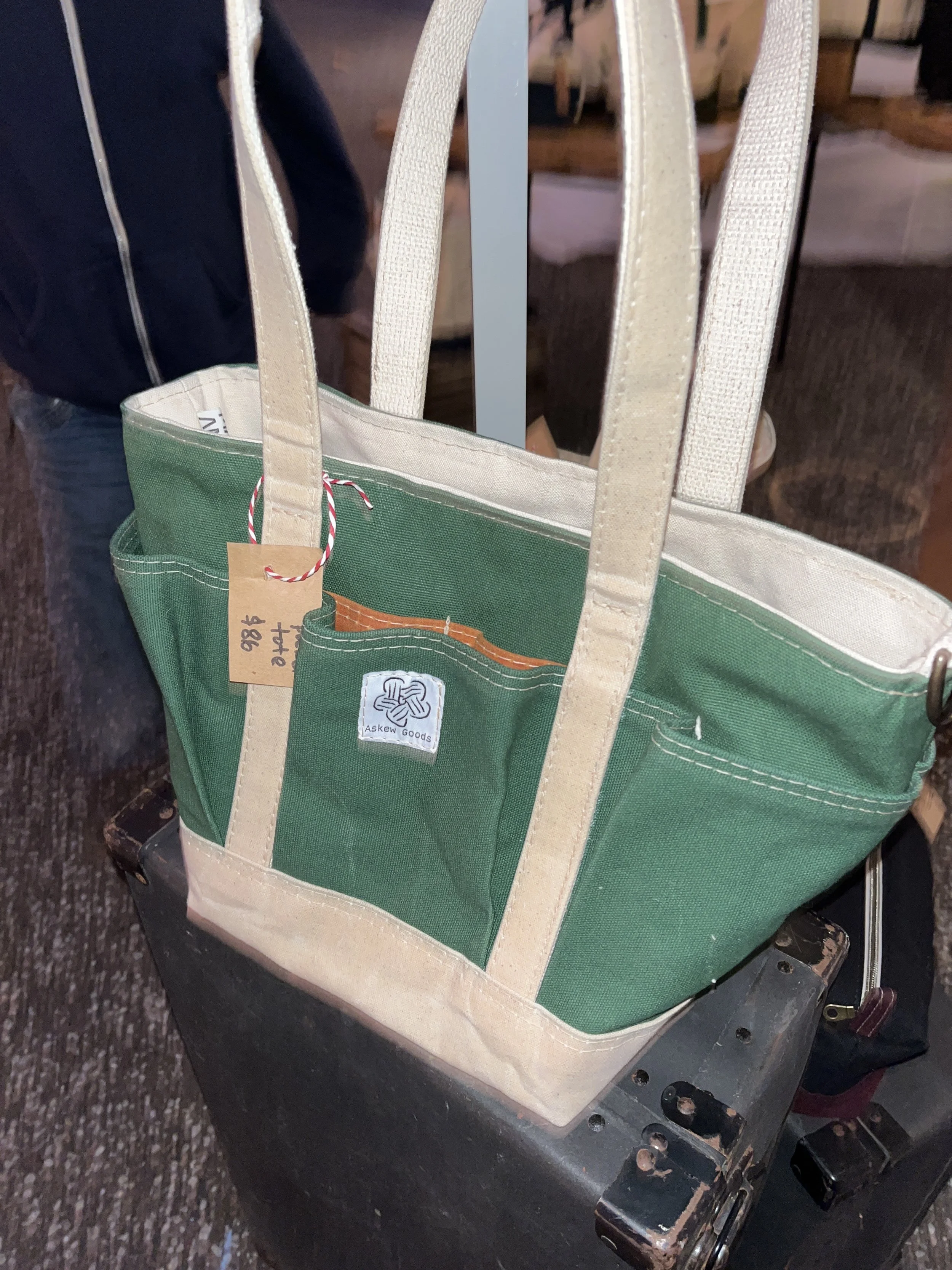 A green and beige tote bag hanging on a luggage cart. The bag has a tag with handwritten text and an Askew Goods logo. The cart is on a dark, textured floor.