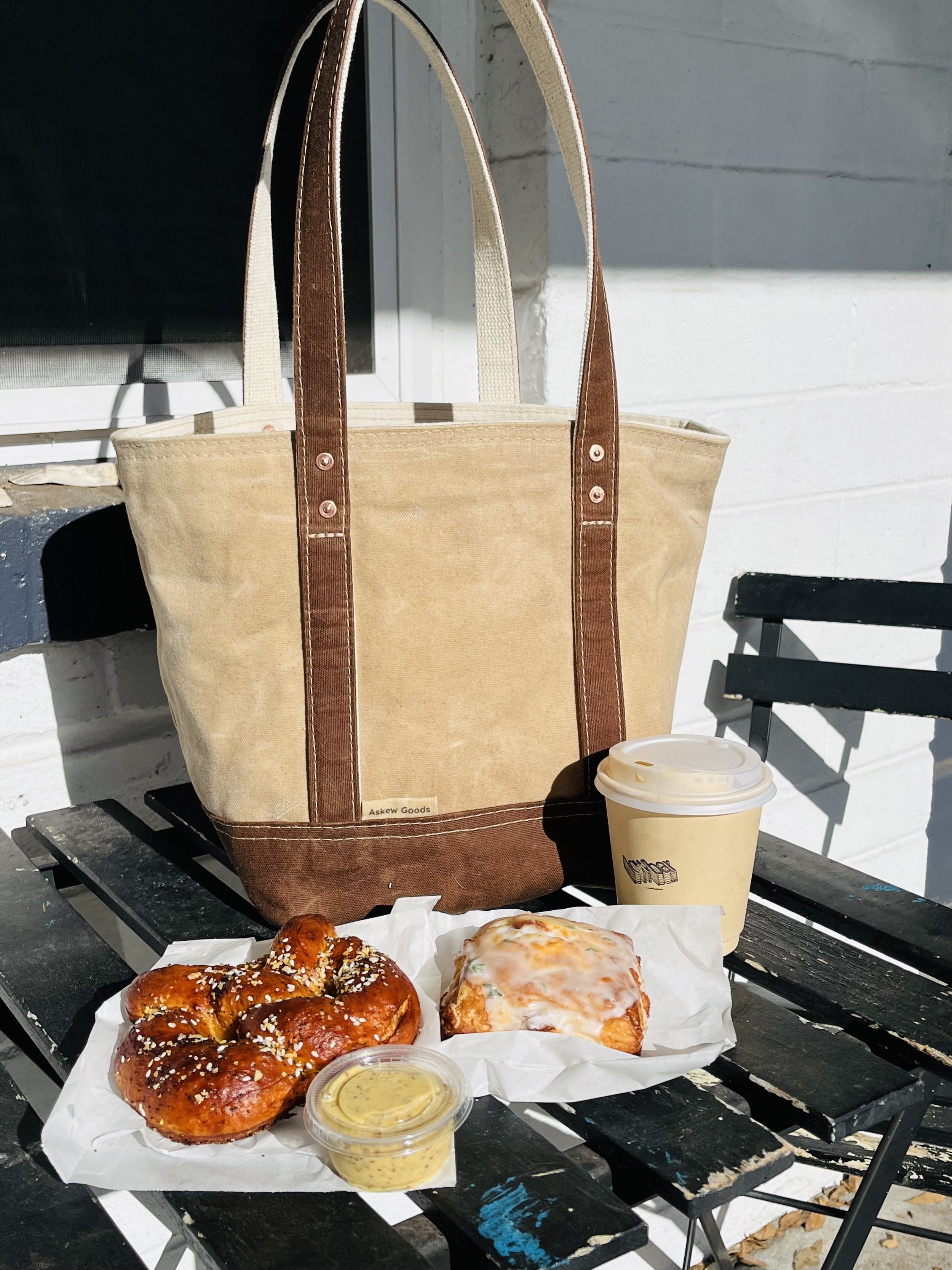 A black picnic table with assorted baked goods like pretzels and a pastry, a small container of mustard, a large beige tote bag with brown handles and a tag that reads 'Askew Goods,' and a paper cup with a plastic lid, set against a white brick wall 