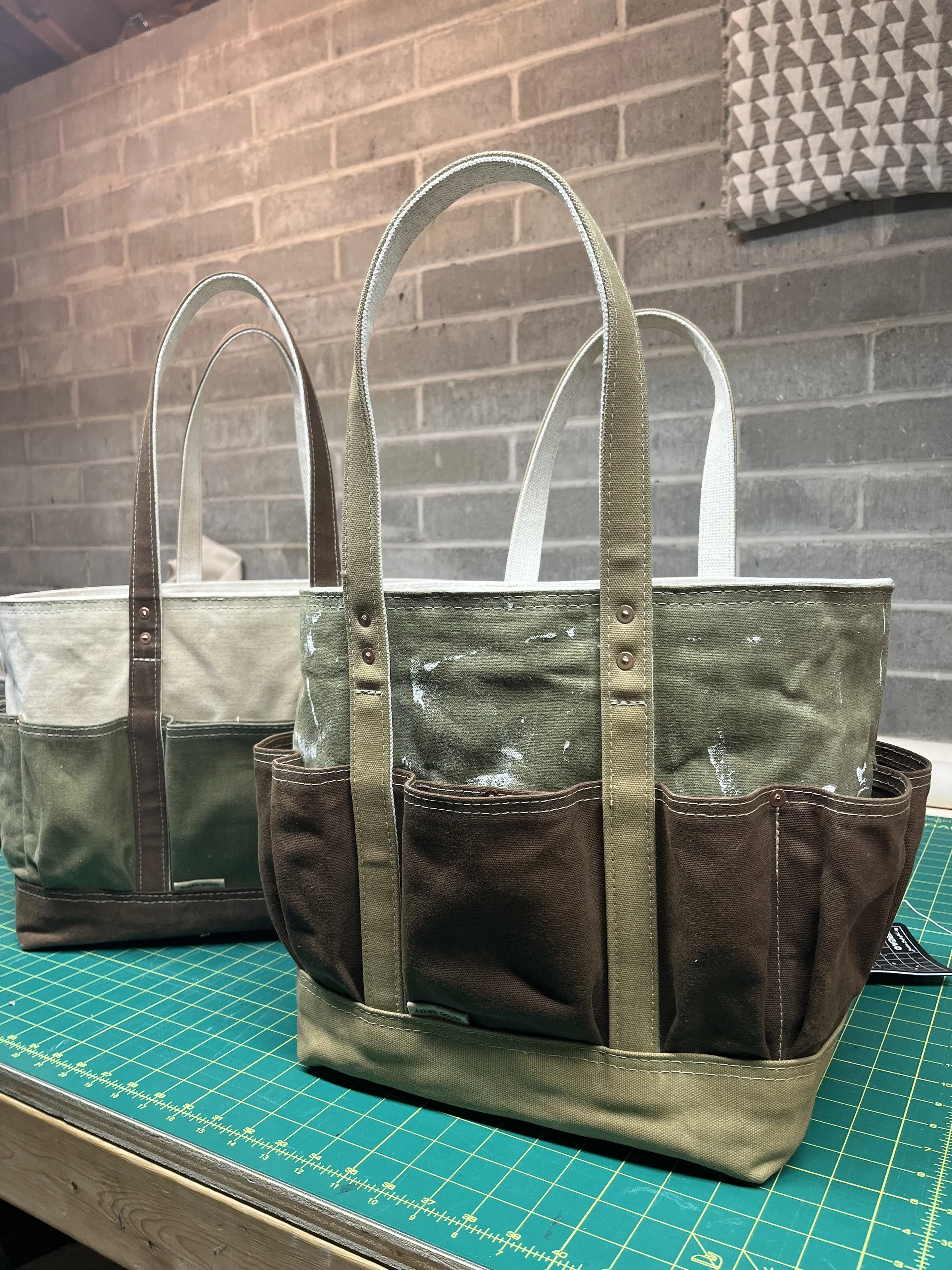 An olive green, brown, and khaki waxed canvas bag sitting upright on a cutting table.