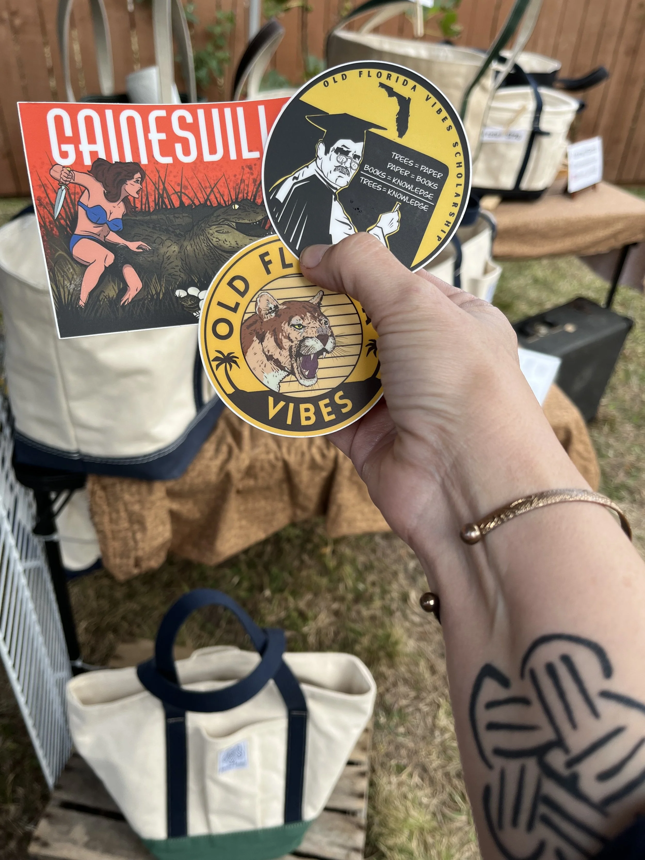 Person holding three stickers with designs and text, at an outdoor market stall with bags and decorative items in the background.