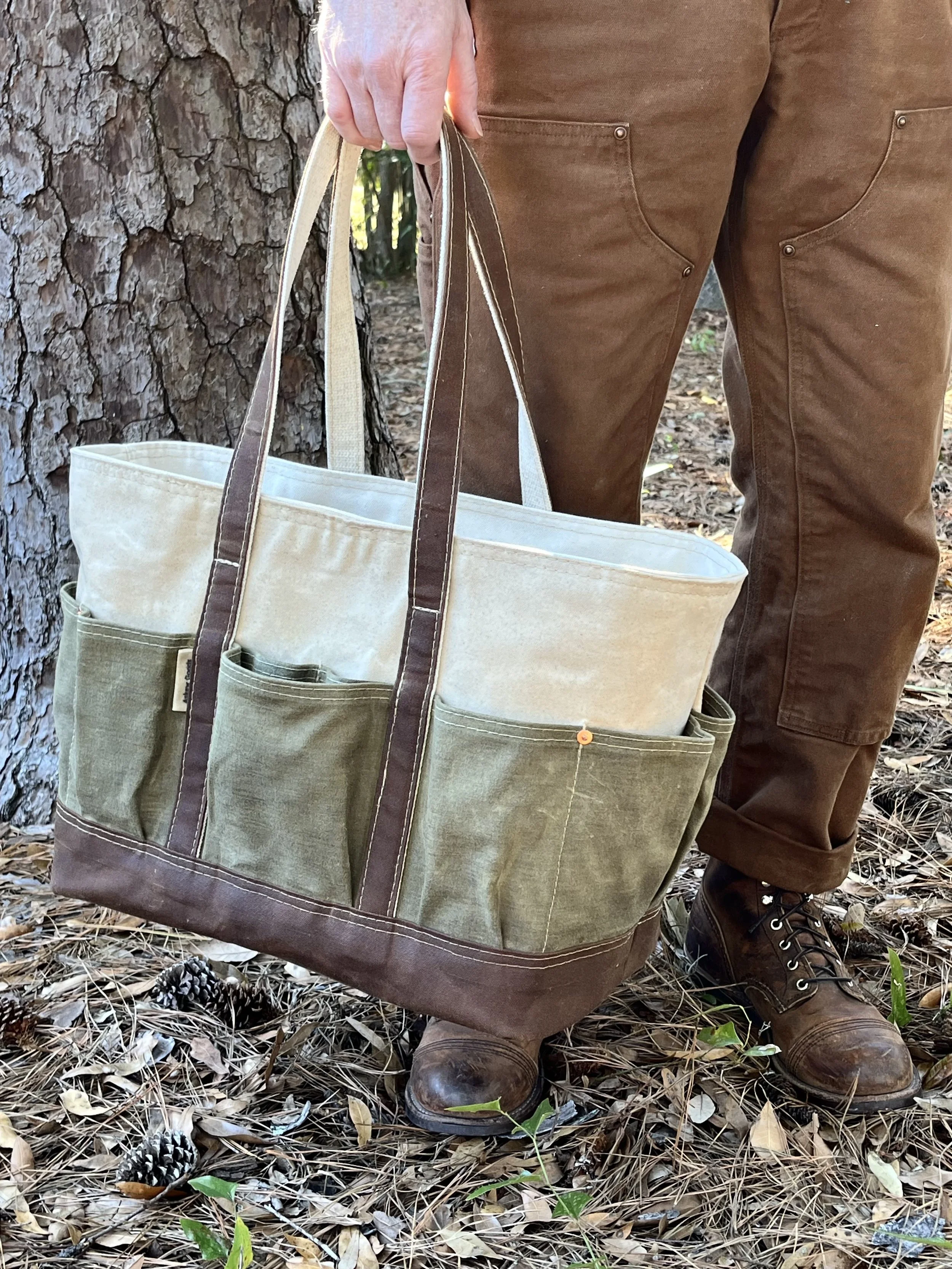 Person wearing brown leather boots and brown pants, holding a beige and green tote bag with large front pockets, standing outdoors on a bed of leaves and twigs near a tree trunk.