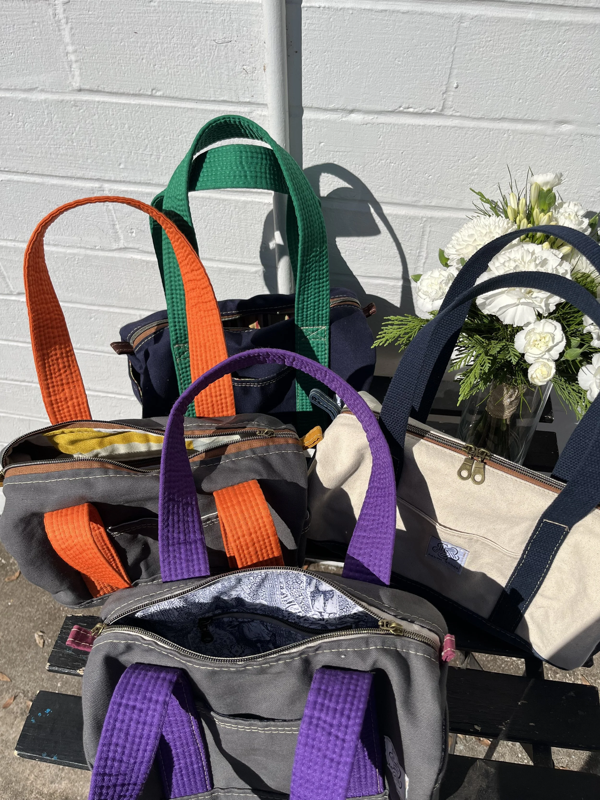 Several backpacks with colorful straps (orange, green, purple, navy, and gray) arranged on a black wooden bench with a white wall and a flower bouquet in a glass vase in the background.