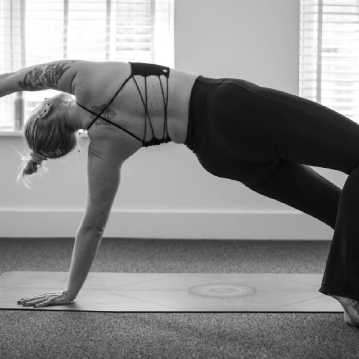 A woman practicing yoga in downward dog pose indoors with large windows and blinds in the background.