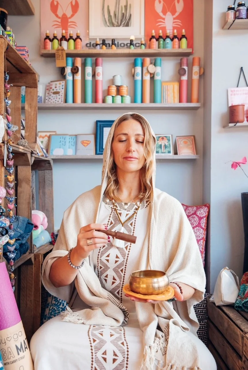 A woman sitting in a colorful shop, holding a singing bowl in one hand and a wooden striker in the other, with her eyes closed. The shop has shelves with various art supplies, framed artwork, and colorful decorations.