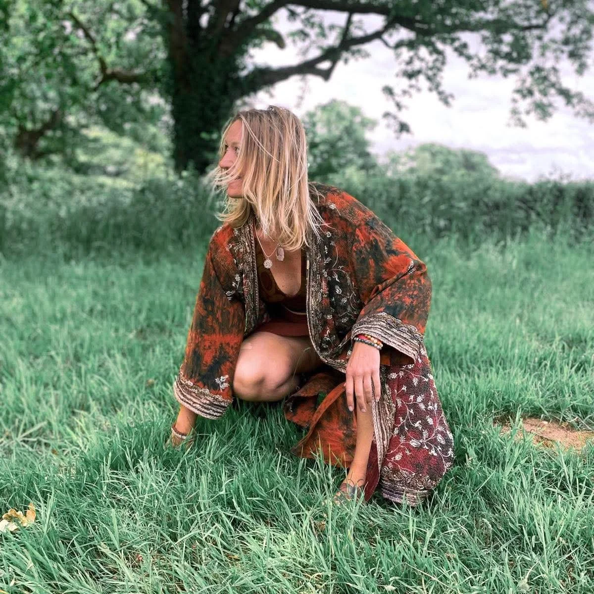 A woman with blonde hair crouches on the grass in a park, wearing a colorful patterned jacket and accessories, with trees and a cloudy sky in the background.