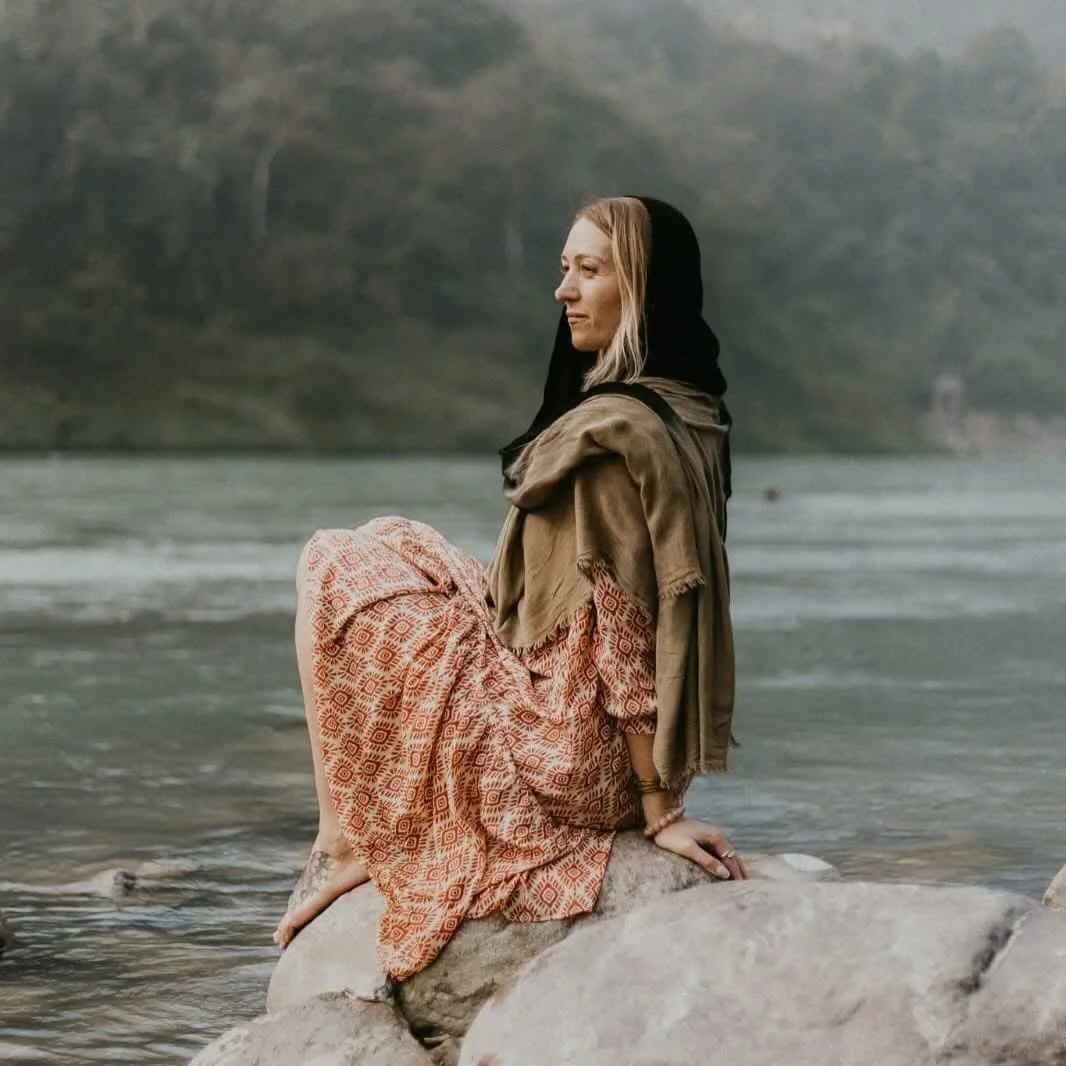 A woman sitting on a rock by a river, dressed in bohemian-style clothing, with a natural background of water and trees.