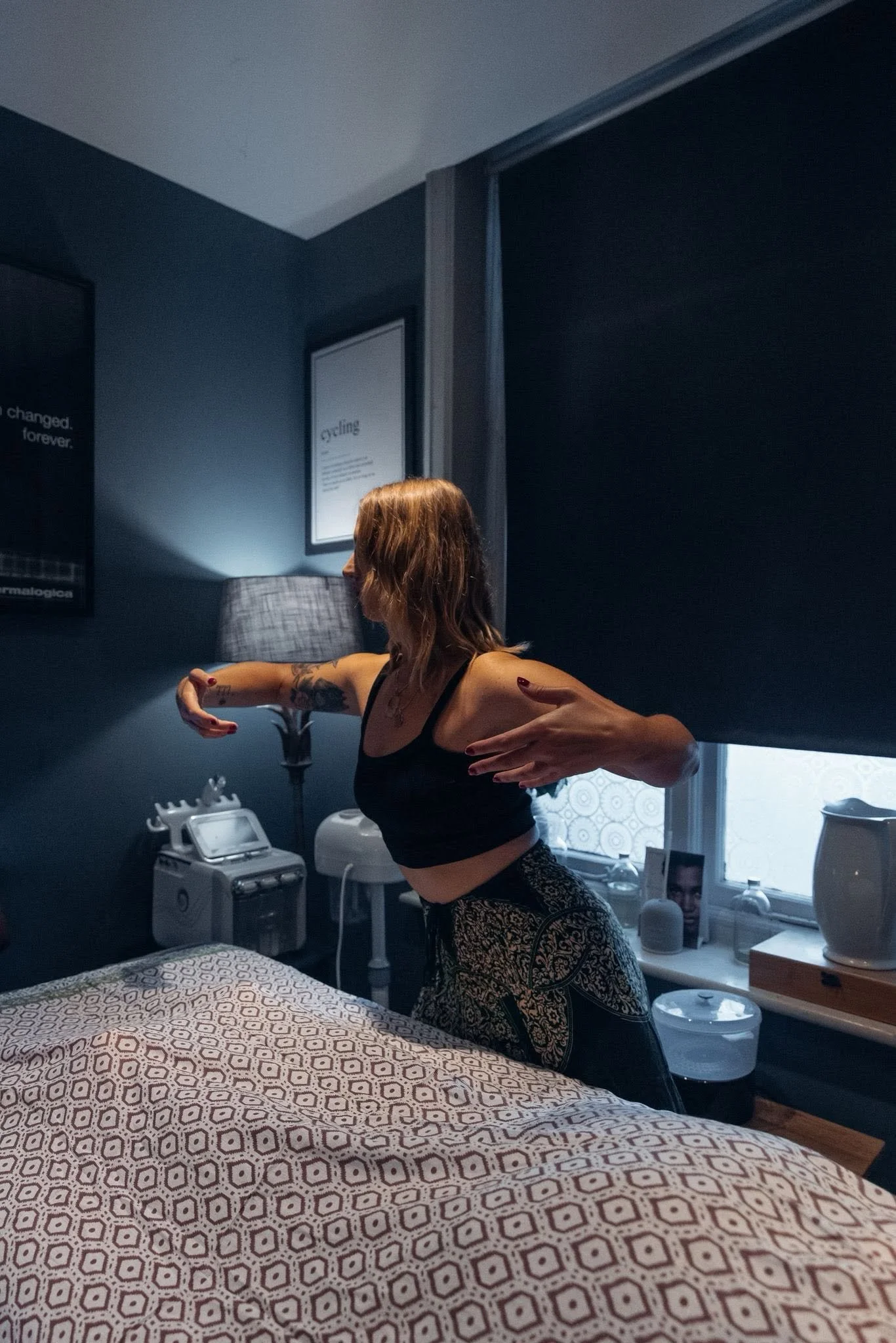 A woman with tattoos on her arms is practicing yoga or stretching in a dimly lit room. She is wearing a black sports bra and patterned pants. The room has a bed with a geometric patterned blanket, a nightstand, a lamp, framed posters on the walls, and various electronic devices on a table.