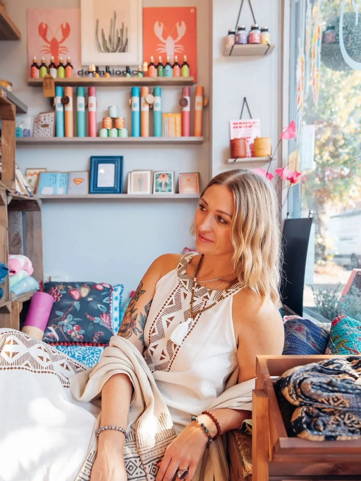 A woman with wavy blonde hair, tattoos on her arm, and wearing a white bohemian dress sitting inside a colorful craft shop or boutique. The shop has shelves with paint bottles, artwork, and decorative items, with a large window letting in natural light.