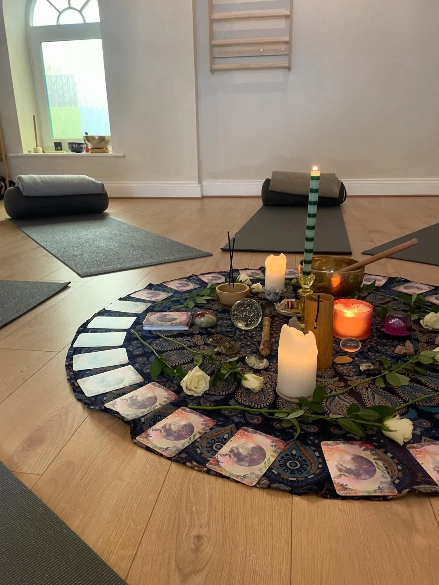 Interior of a room with yoga mats, a decorated circular cloth on the floor with candles, crystals, and tarot cards, and a wall with a window and wall hanging.