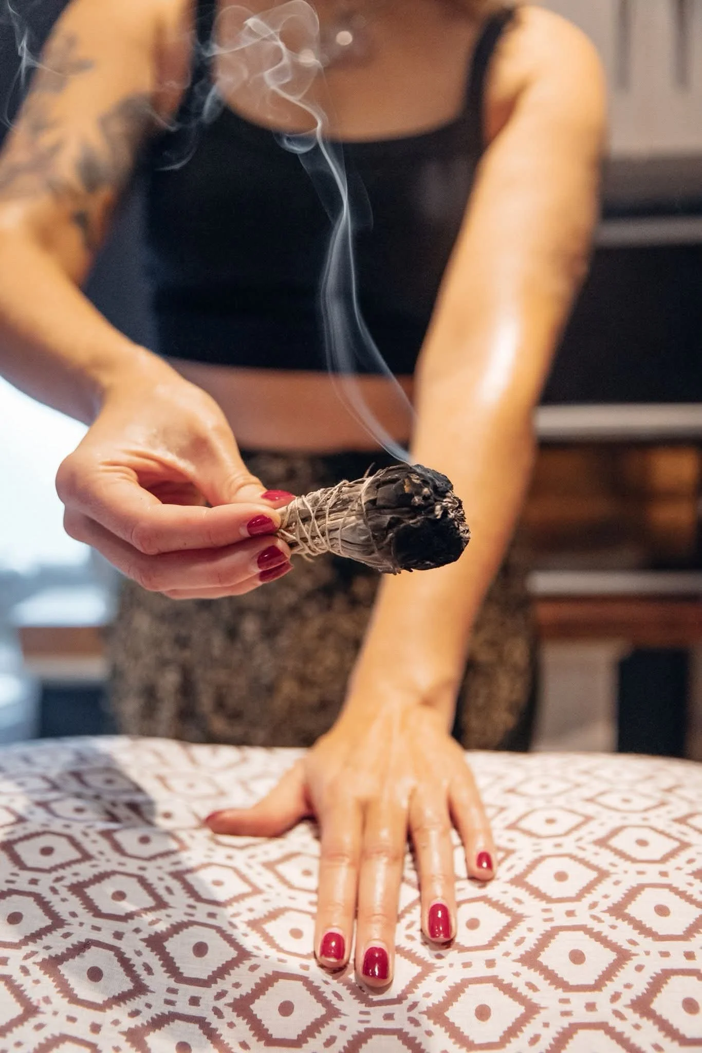 A person holding a smoldering bundle of sage, with smoke rising, while their hand and fingers are resting on a table covered with patterned fabric. The person is wearing a black sleeveless top, and has tattoos on their arms. Their nails are painted red.