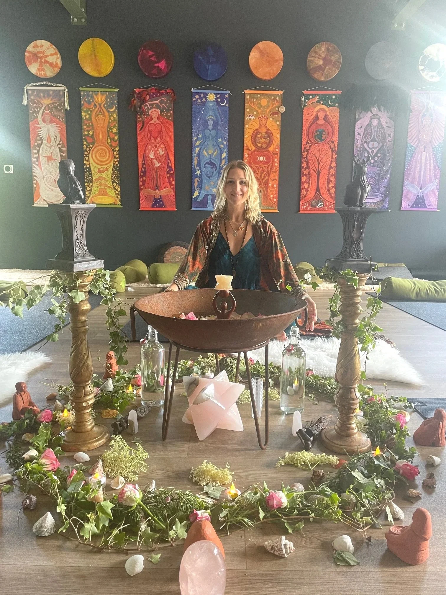 A woman smiling sitting at a table decorated with flowers, candles, and shells, with colorful tapestries hanging on a dark wall behind her in a spiritual or ceremonial setting.