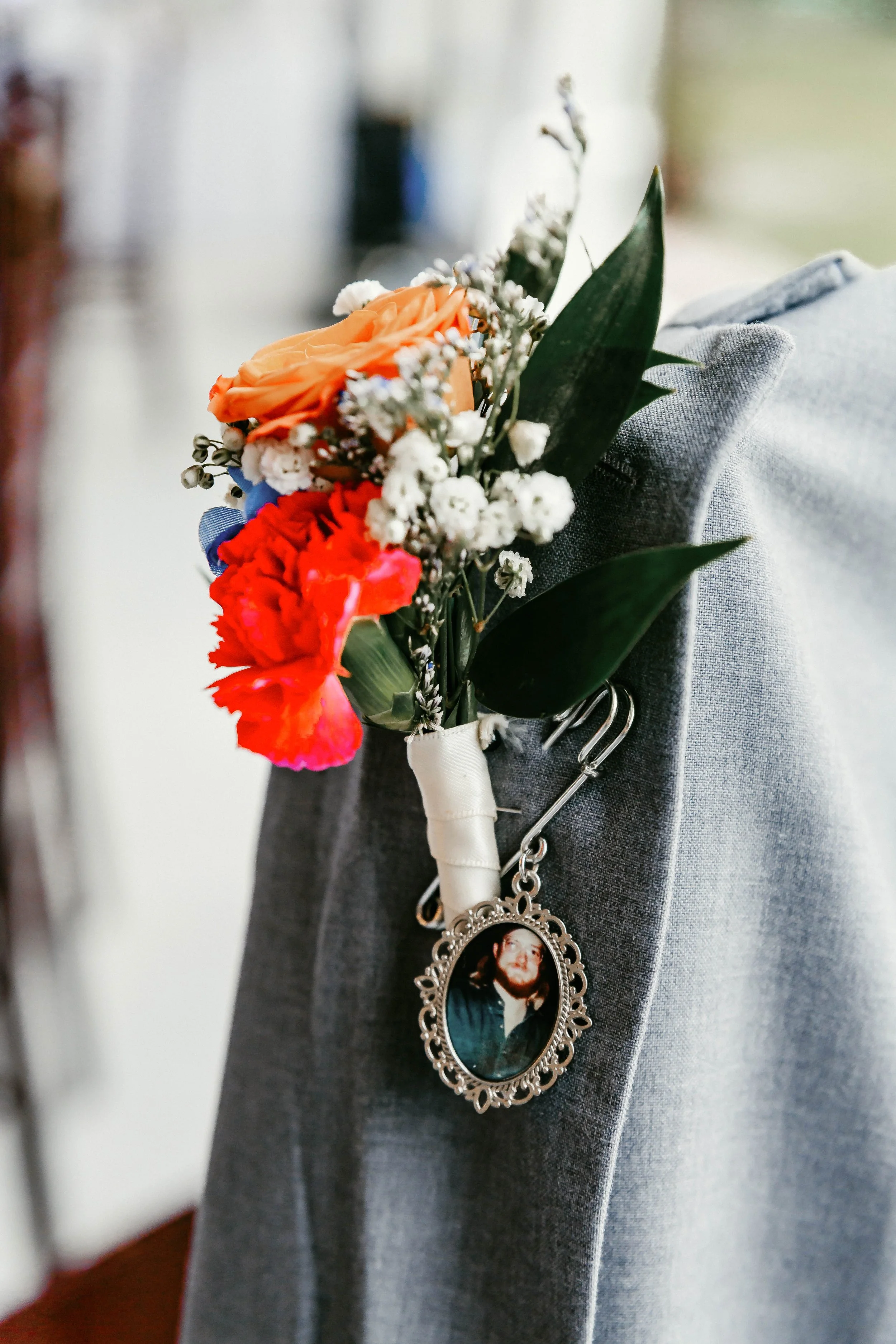 A boutonniere with peach, red, and white flowers pinned to a gray fabric surface, featuring a medical alert charm with a photo of a woman.