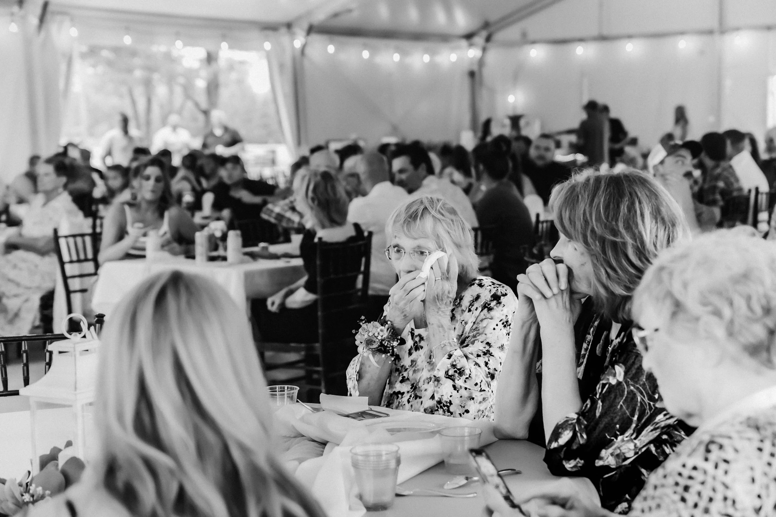 A black and white photo of a large gathering of people inside a decorated tent, seated at tables, possibly at a wedding or celebration. In the foreground, two women appear emotional, one wiping her eye with a tissue, while the other has her hands cla