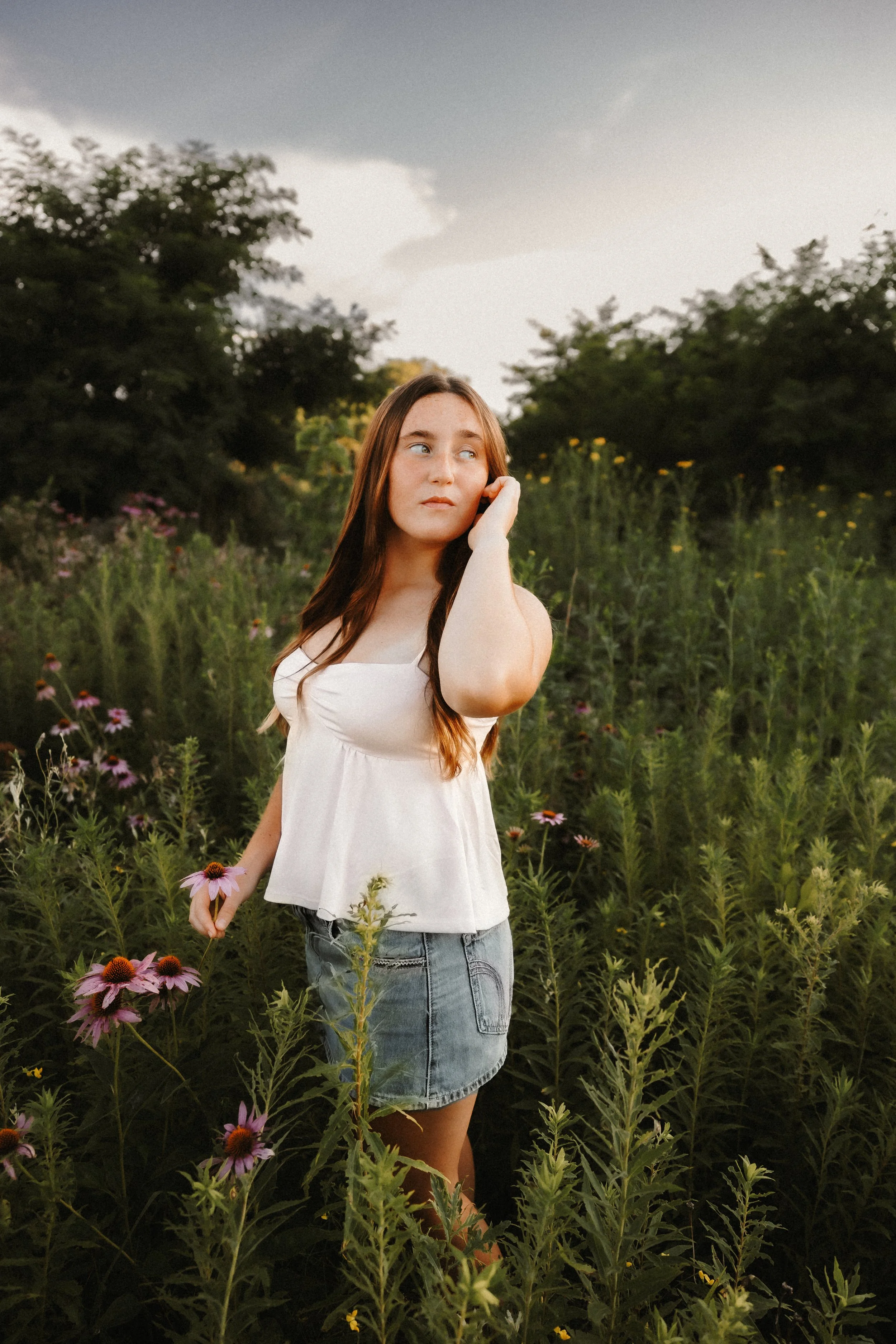 A young woman with long brown hair, wearing a white sleeveless top and denim skirt, standing in a field of wildflowers with a cloudy sky in the background.