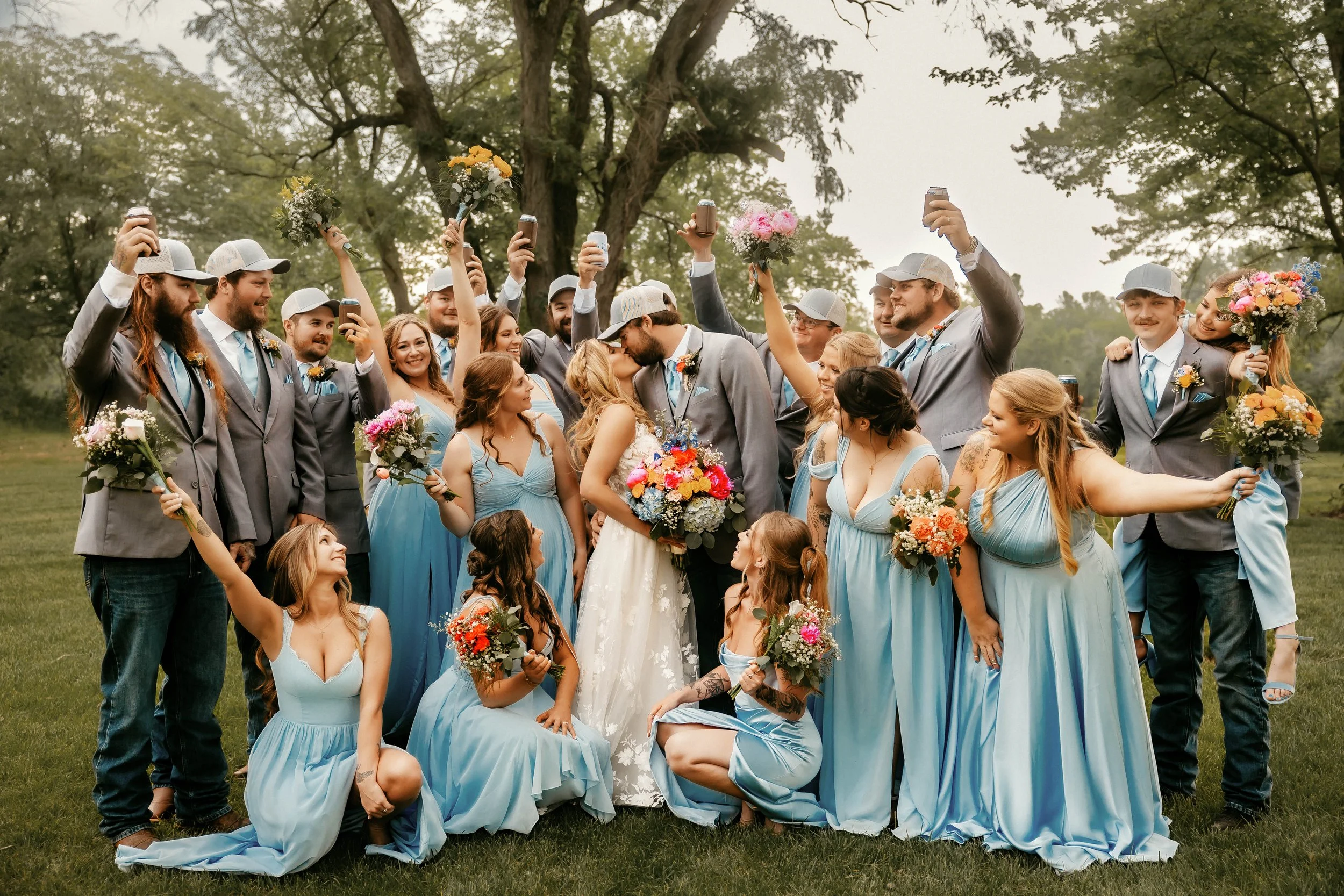 A large group of wedding guests, wearing gray suits and light blue dresses, celebrating outdoors around a bride and groom who are kissing. The guests are holding flowers and taking photos, with some raising their phones and drinks in the air, on a gr