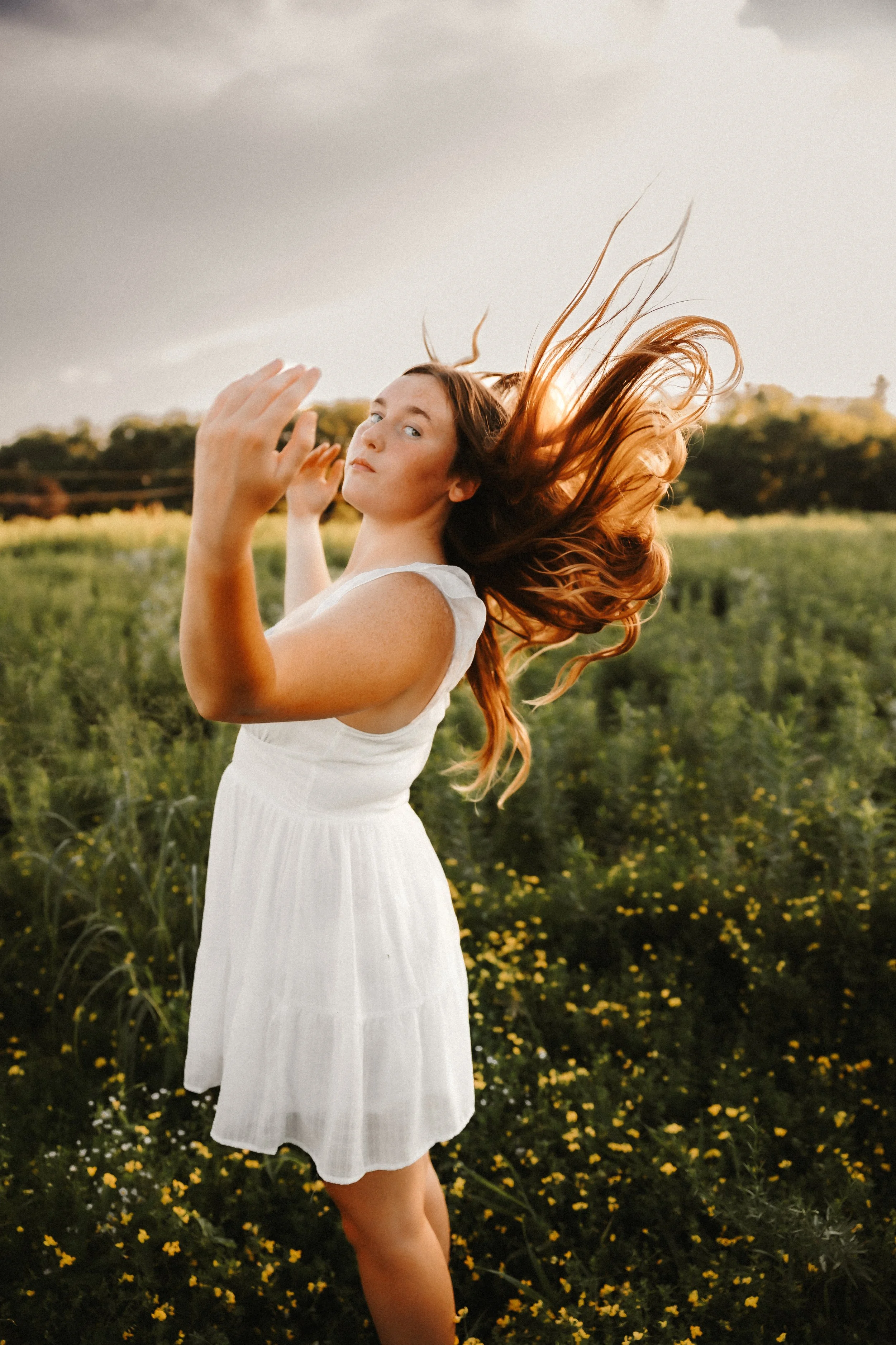A woman with long, flowing red hair in a white dress standing in a field of yellow flowers during sunset or late afternoon.