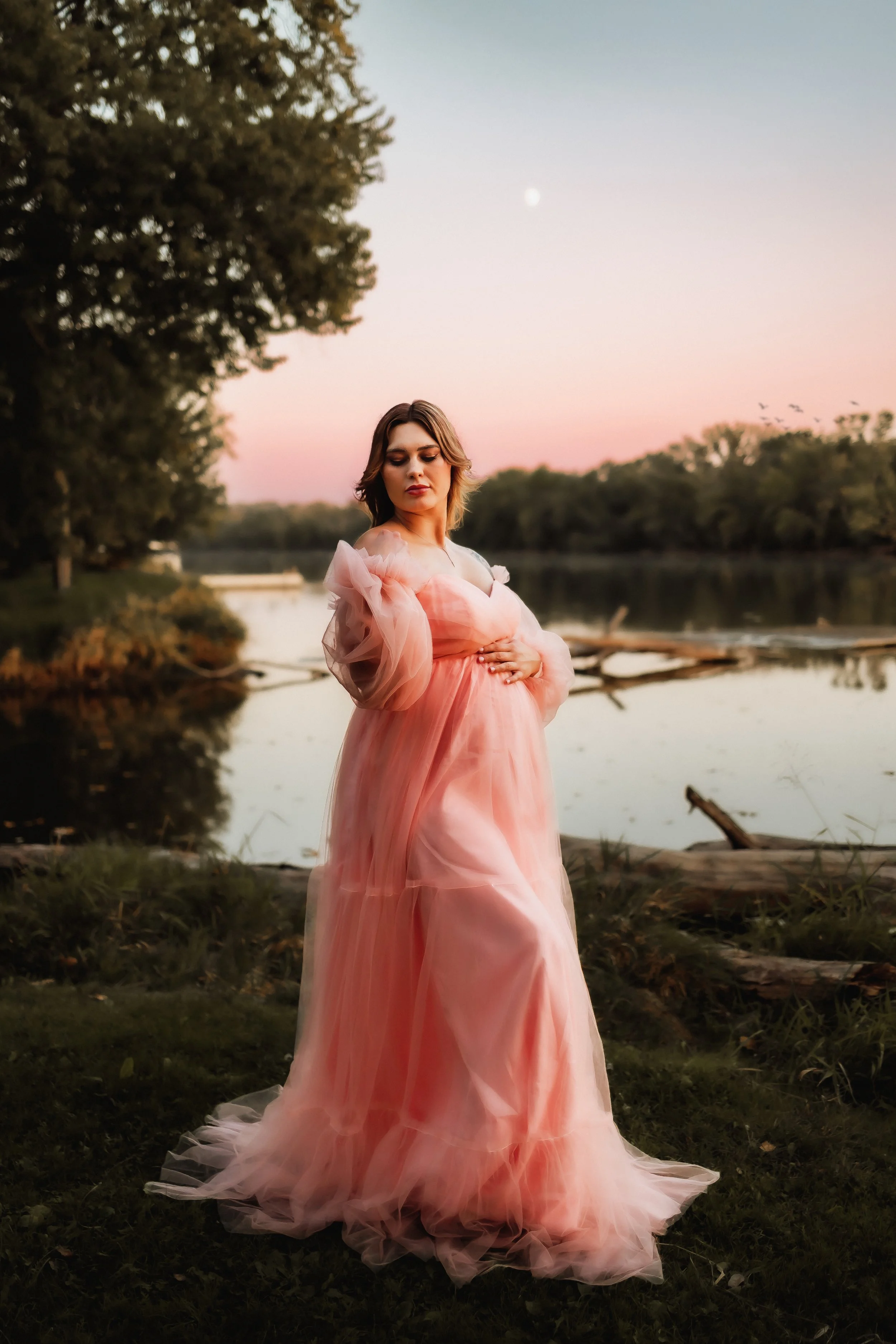A pregnant woman in a flowing pink dress stands near a river at sunset, hand on her belly.