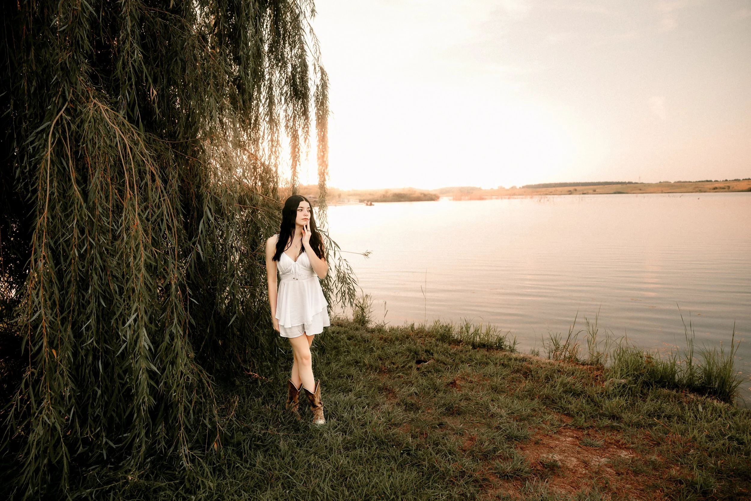 A woman in a white dress and cowboy boots stands next to a tree by a lake at sunset, looking contemplatively into the distance.