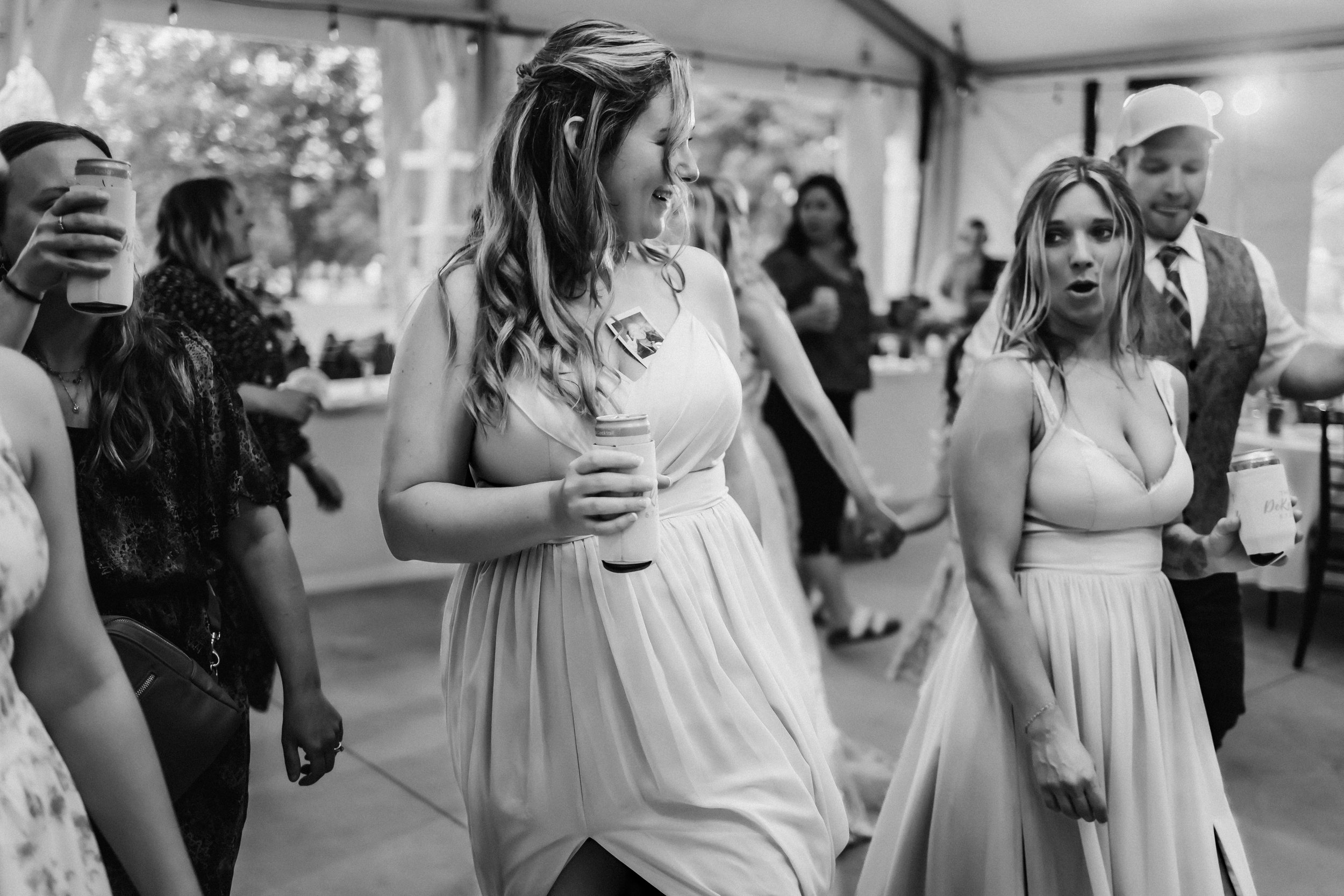 People dancing at a wedding reception inside a tent, both women wearing light-colored dresses and holding drinks, with other guests in the background.