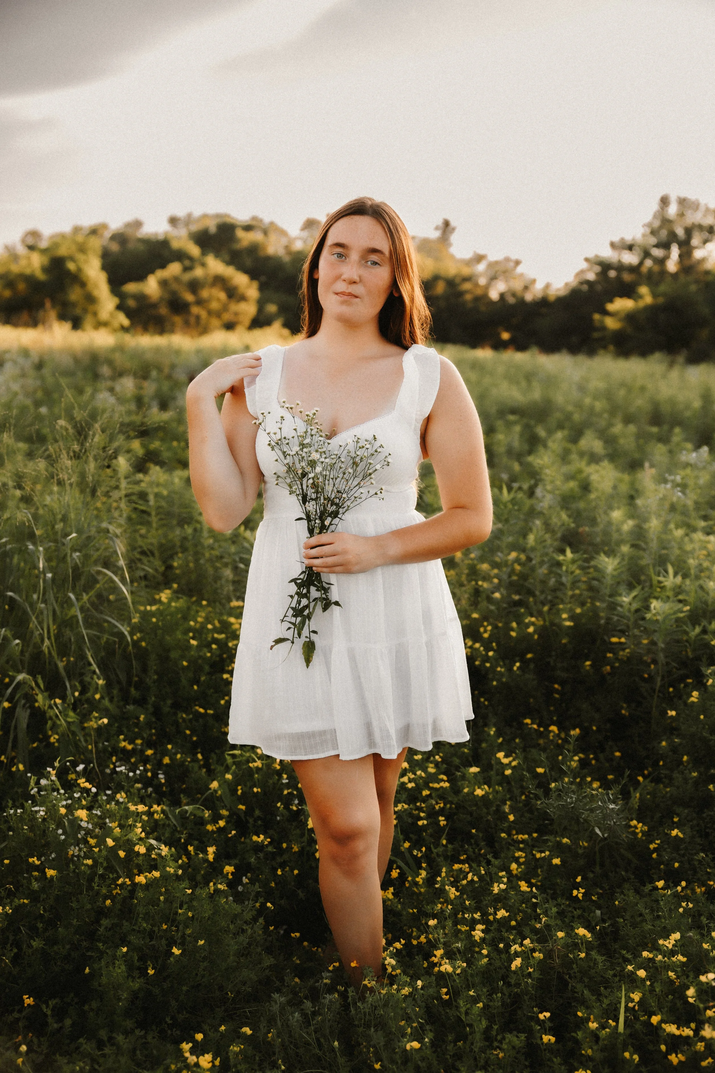 A young woman in a white dress standing in a green field holding a small bouquet of white flowers, with trees and a cloudy sky in the background.