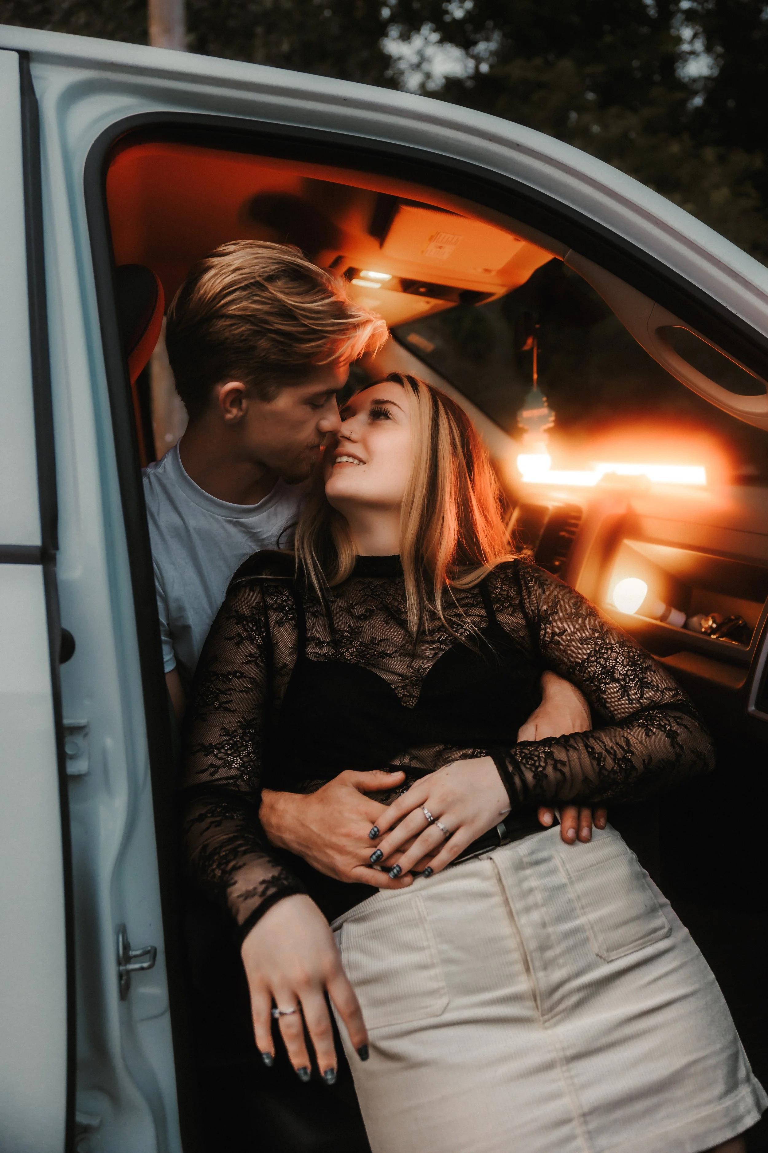 A young couple sharing an intimate moment inside a car at sunset, with the man leaning in towards the woman who is smiling while sitting in the passenger seat.