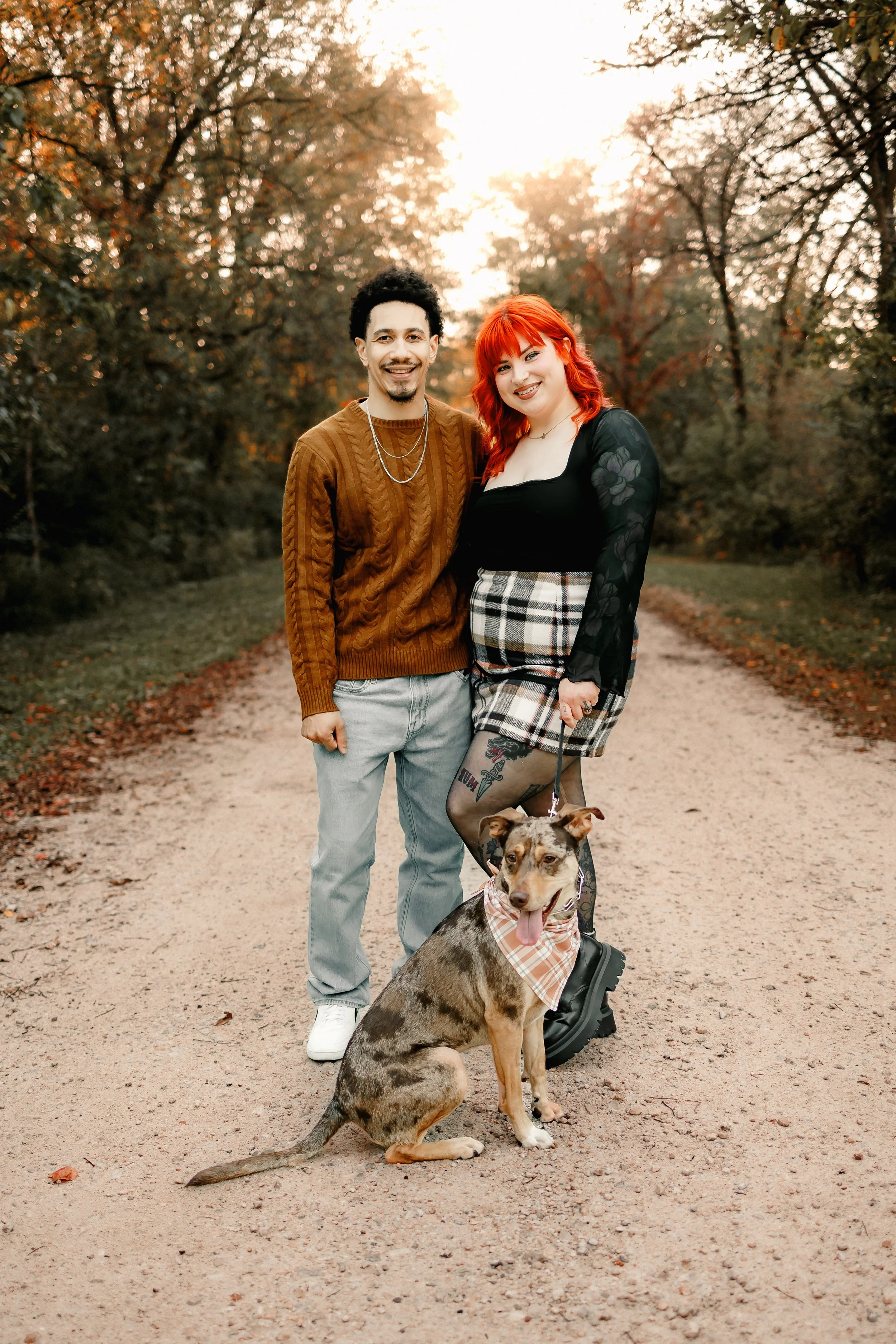 A smiling man and woman stand on a dirt path in a wooded area during sunset, with a dog sitting in front of them, all enjoying an autumn walk.