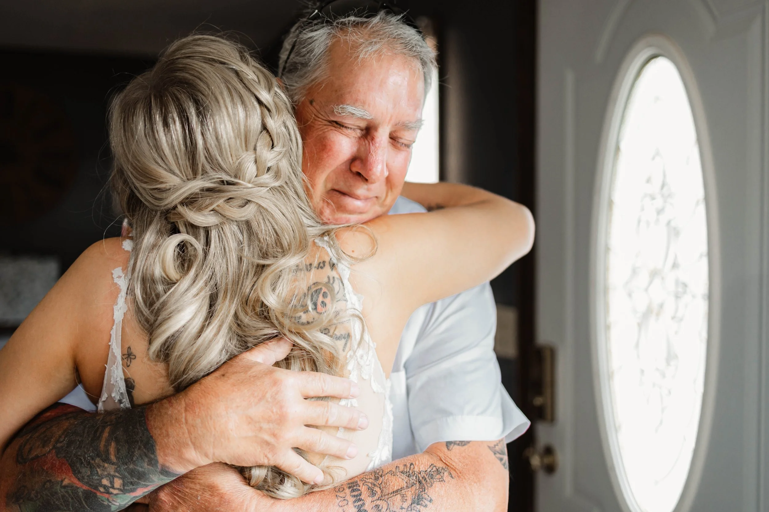 An elderly man and a woman with long curly hair hugging each other tightly inside near a door with glass panels.